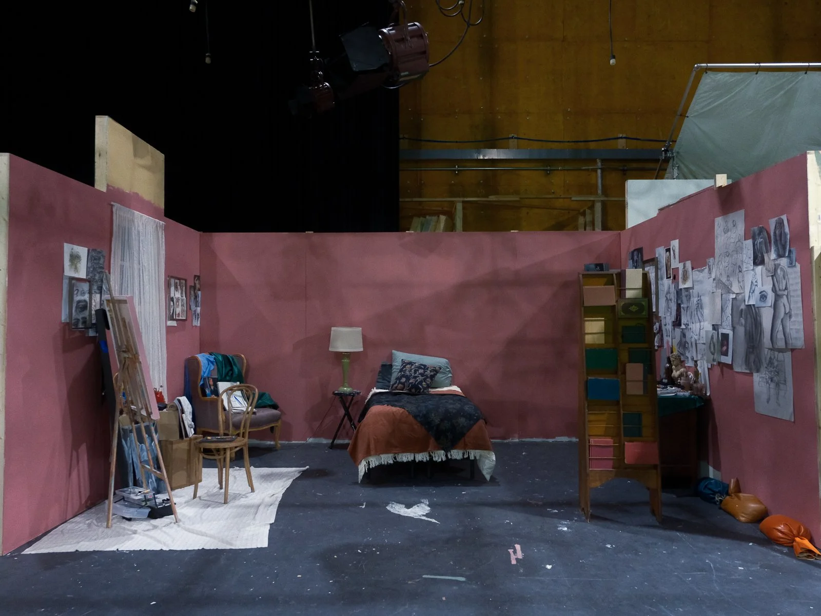 A small interior set resembling a bedroom with pink walls, a bed with an orange blanket, a lamp on a small table, artwork on the walls, and various furniture and art supplies scattered around.