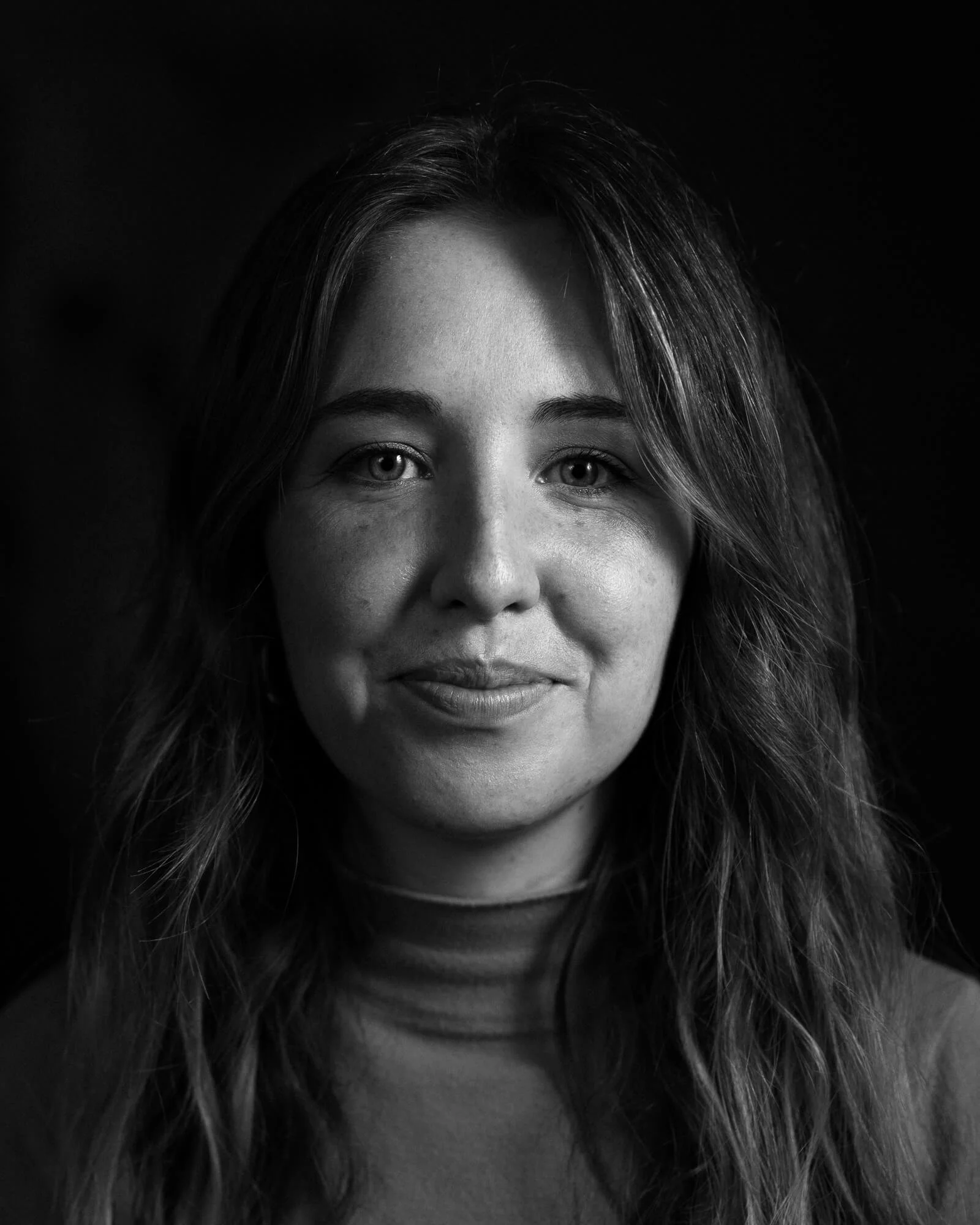 A black and white portrait of a woman with long, wavy hair and light-colored eyes, smiling softly at the camera.