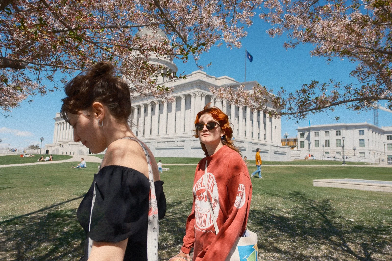Two women standing outdoors under blooming cherry blossom trees with the Washington State Capitol building in the background.