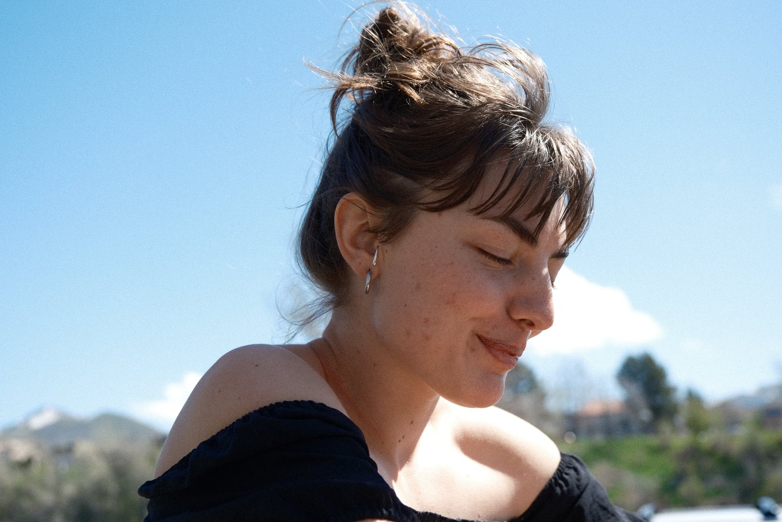 A young woman with short brown hair and earrings, smiling with eyes closed, outdoors on a sunny day.
