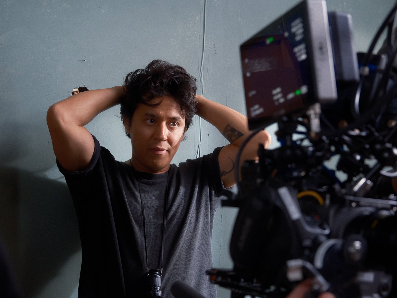A man with curly dark hair, wearing a black t-shirt, stands against a plain wall with wires behind him. He is looking at a camera setup with intense focus, and has his hands behind his head, adjusting his hair.