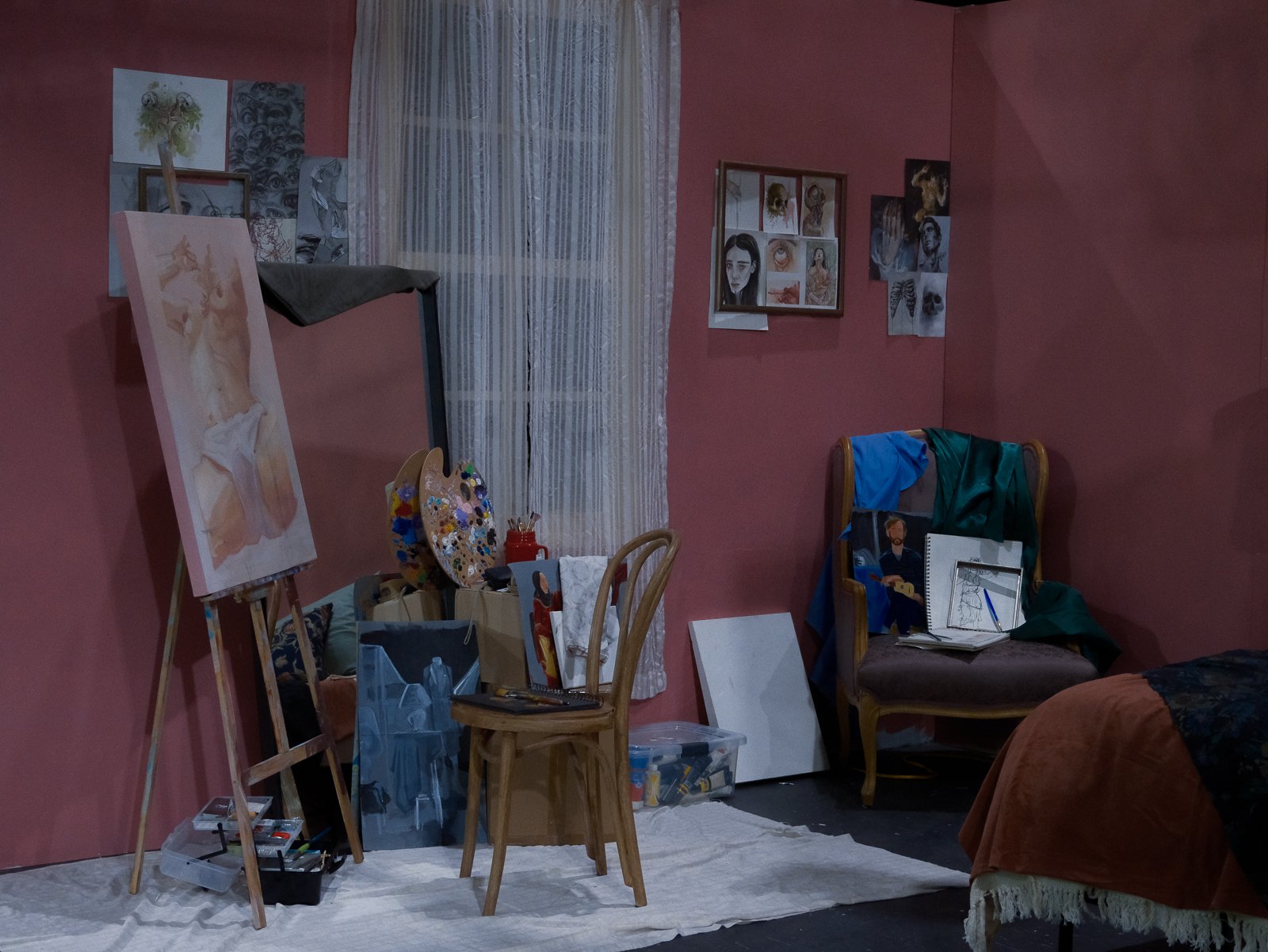 Art studio with pink walls, featuring a painting on an easel, a window with sheer curtains, framed artwork on the walls, and chairs with art supplies and clothes.