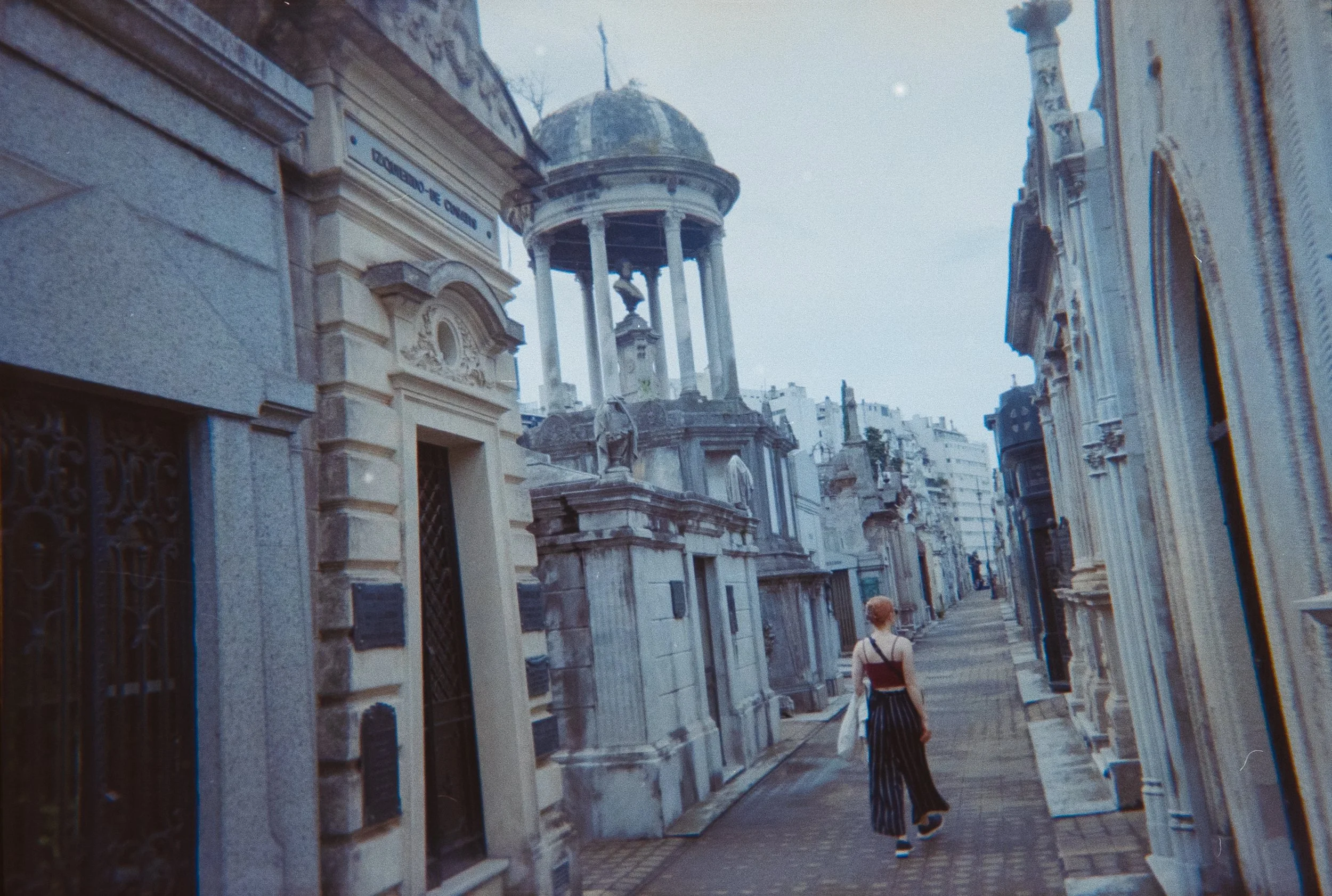 A city street with historic stone buildings and ornate architectural details. A person with a black top and striped pants walks along the sidewalk. The sky appears overcast.
