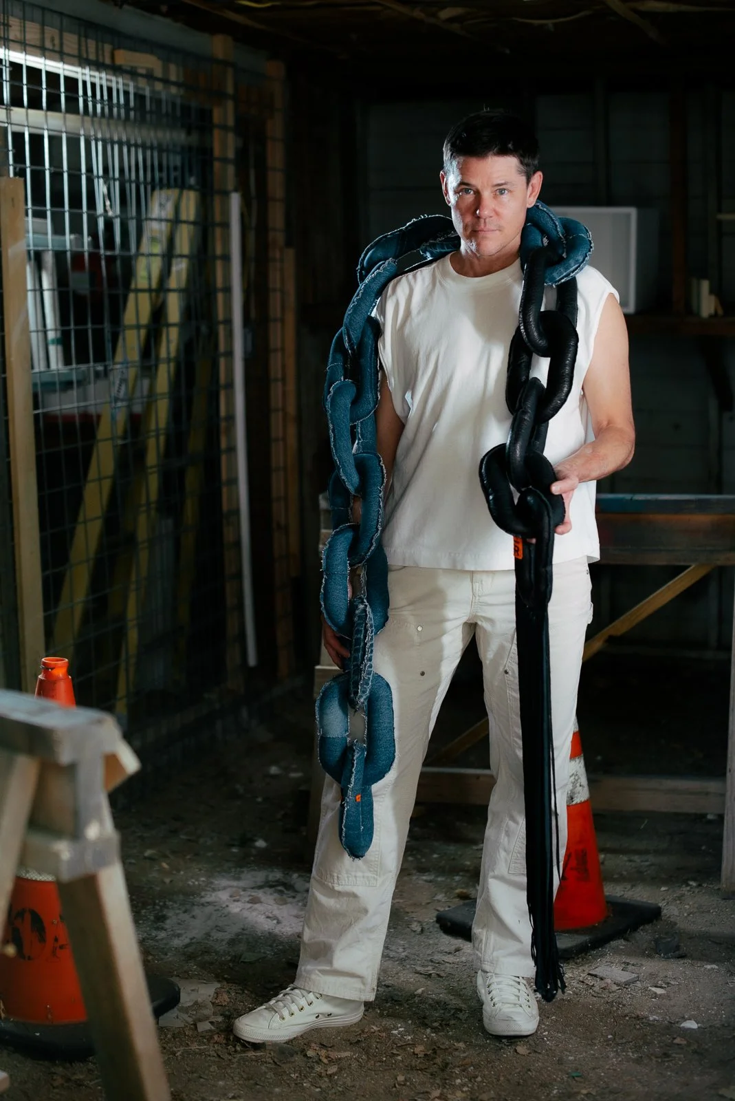 A man in a white sleeveless shirt and white pants is standing in a warehouse or garage, holding a large snake made of fabric or leather. The snake is draped around his shoulders and hanging down in front of him.
