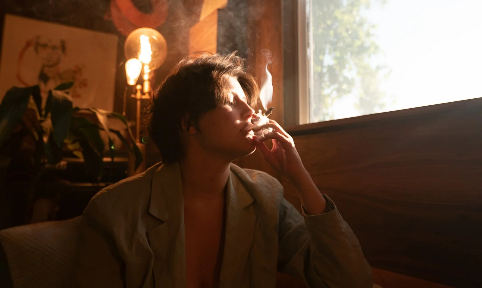 A person with short dark hair smoking a cigarette by a window with sunlight coming in, inside a cozy room with a lamp, a painting, and a plant in the background.