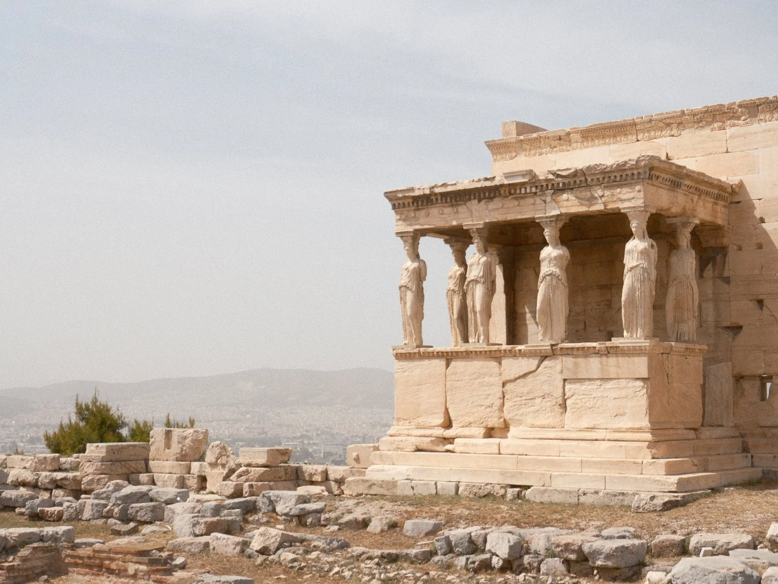 Ancient Greek temple with six standing columns and detailed carvings, ruins scattered in front, against a backdrop of distant hills and a partly cloudy sky.
