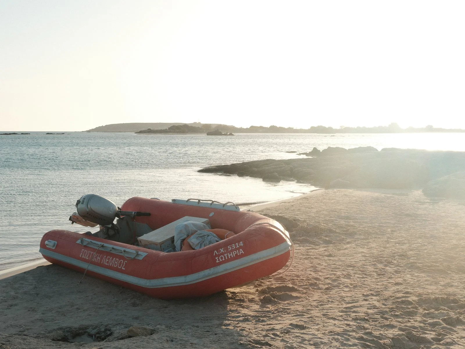 An orange rescue boat resting on a sandy beach near the water, with a distant island and rocks under a bright, overexposed sky.