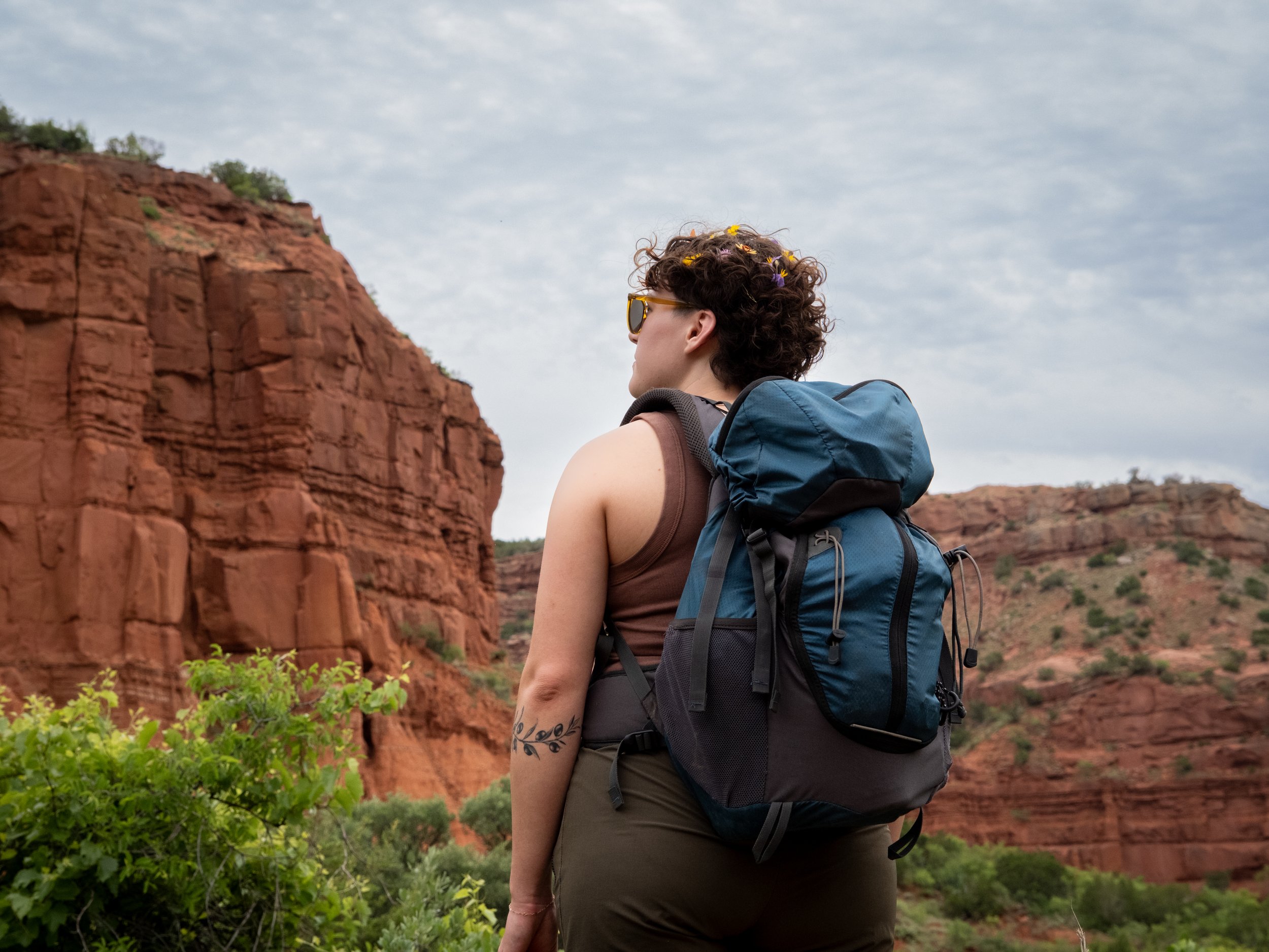 A woman with curly hair and sunglasses carrying a blue backpack, hiking in a desert landscape with red rock formations and a cloudy sky.