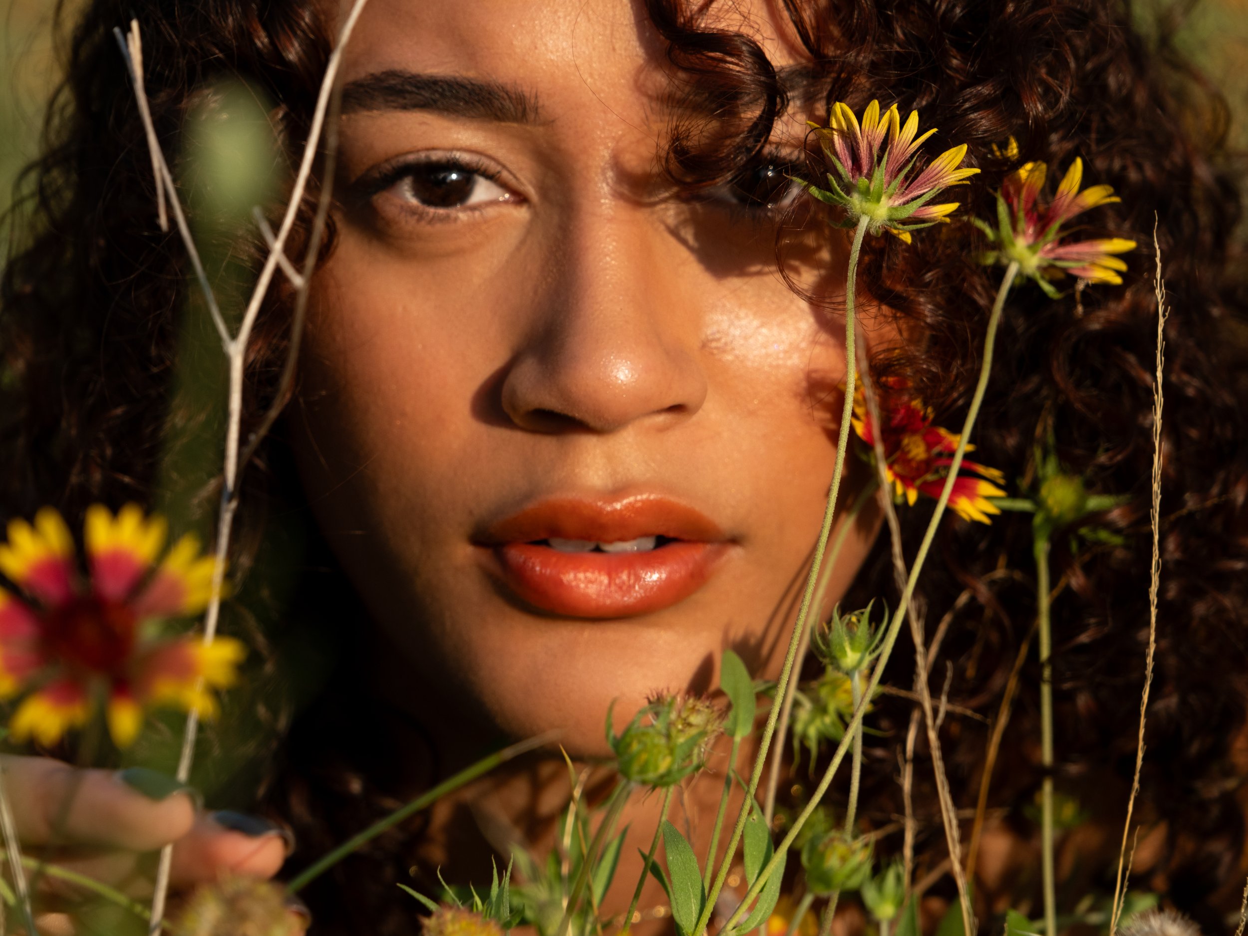 A close-up of a woman with curly hair partially obscured by yellow and reddish flowers, with her eye and part of her face visible.