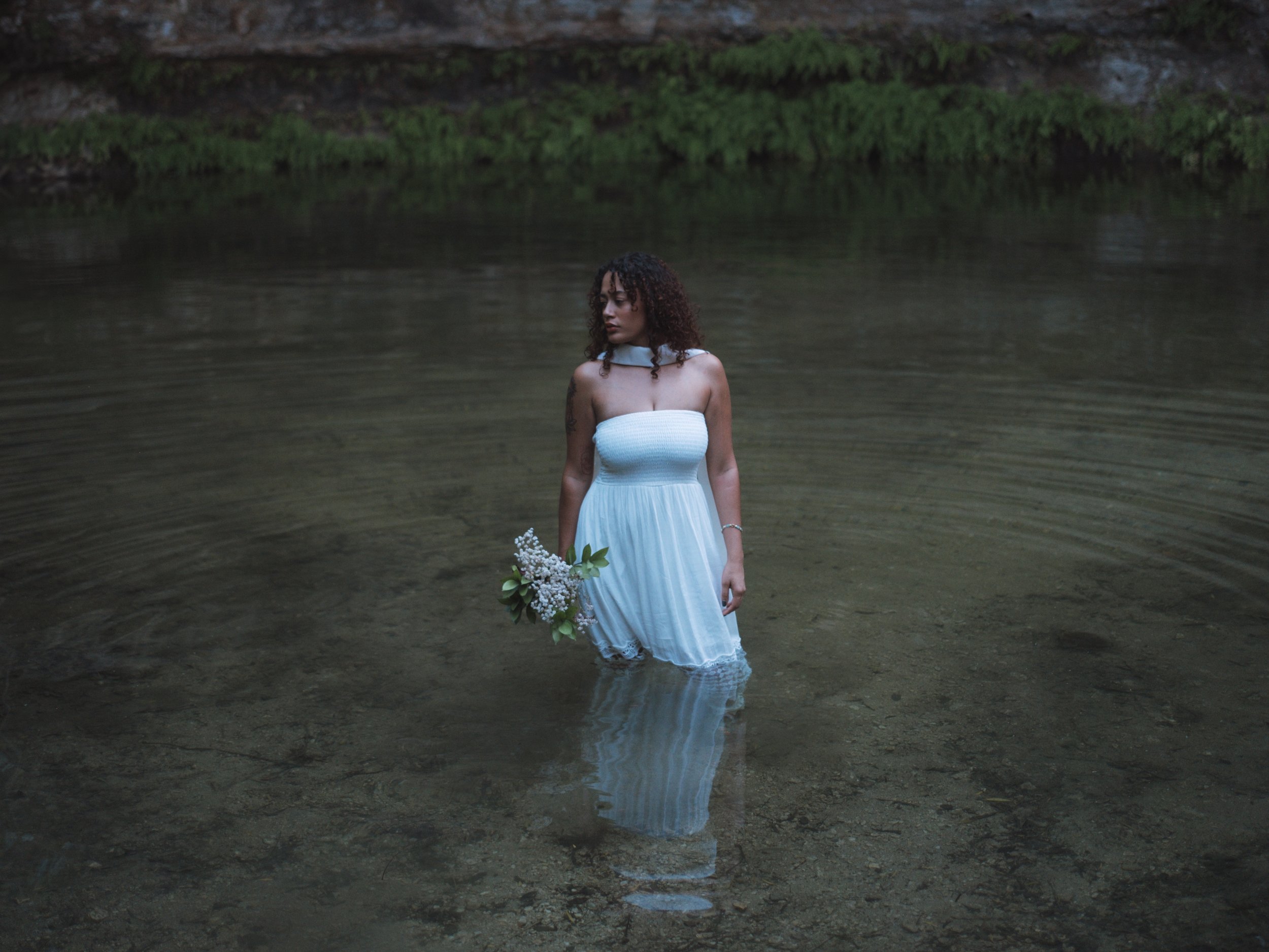 A woman wearing a white strapless dress standing in a body of water, holding a bouquet of white flowers with green leaves, with a natural landscape in the background.