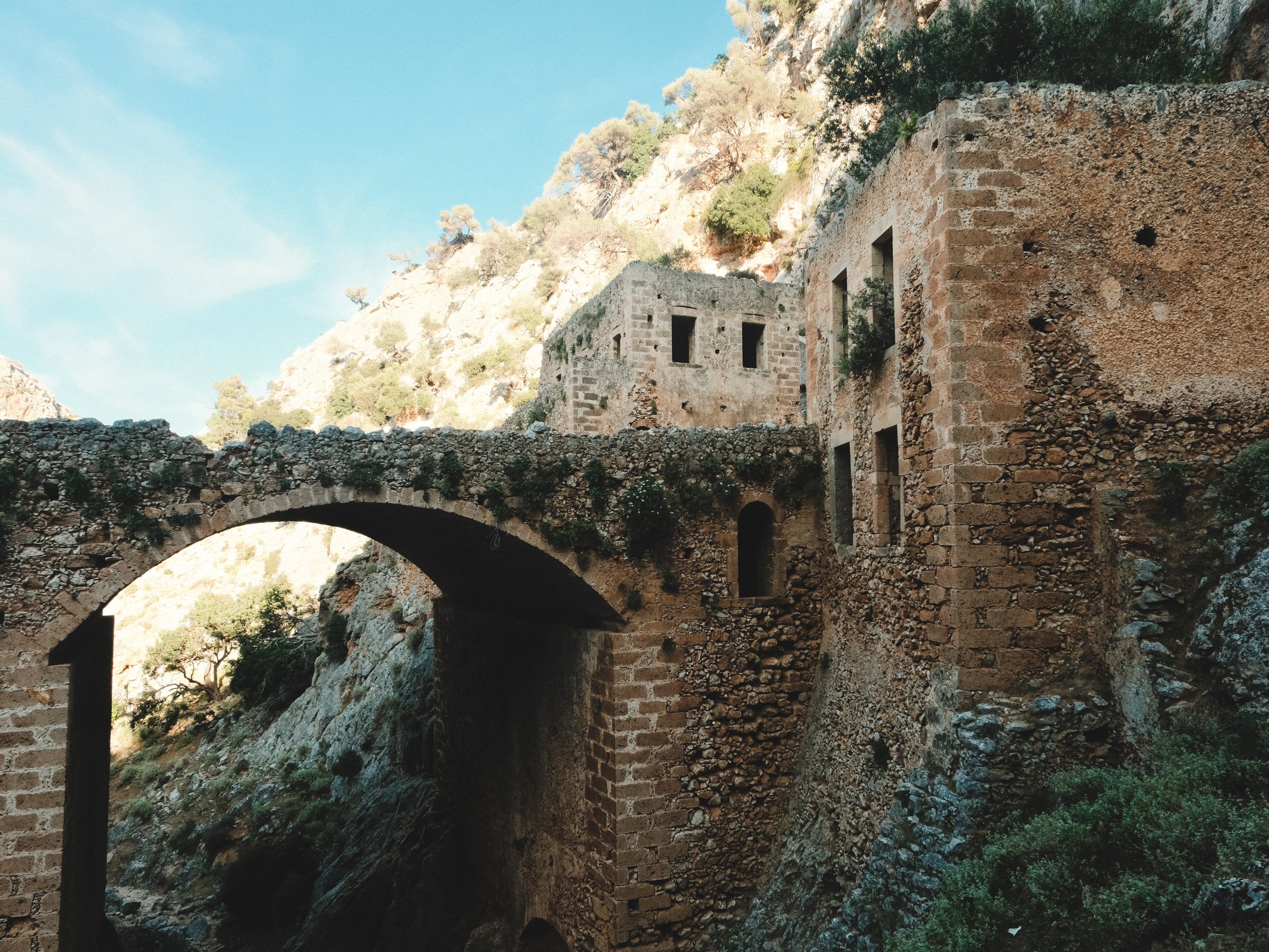 An old stone building with multiple windows, situated on rocky terrain next to a stone bridge, with trees and a mountain hillside in the background under a clear blue sky.