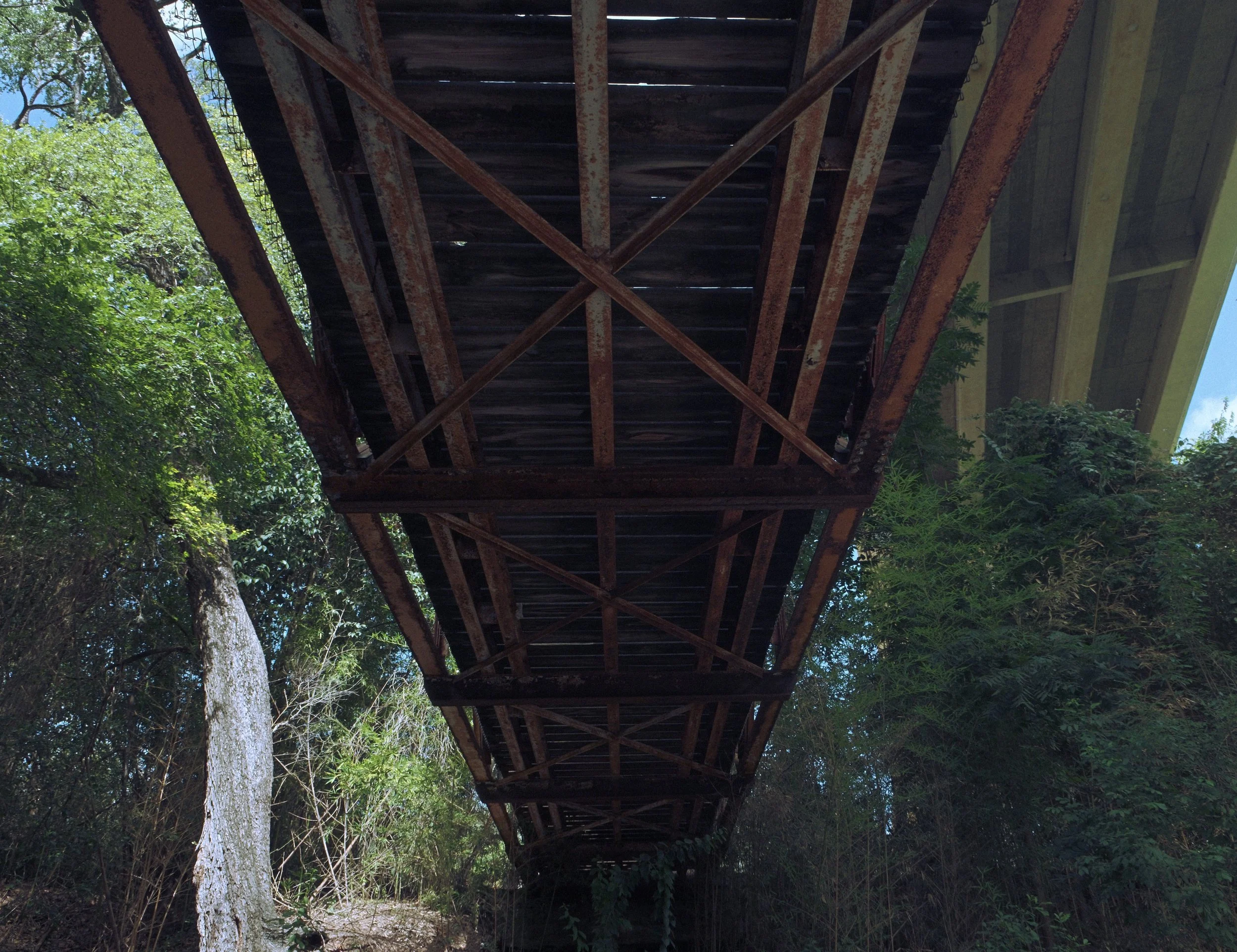 Looking up at the underside of a rusty metal bridge with wooden planks, surrounded by green trees and blue sky.