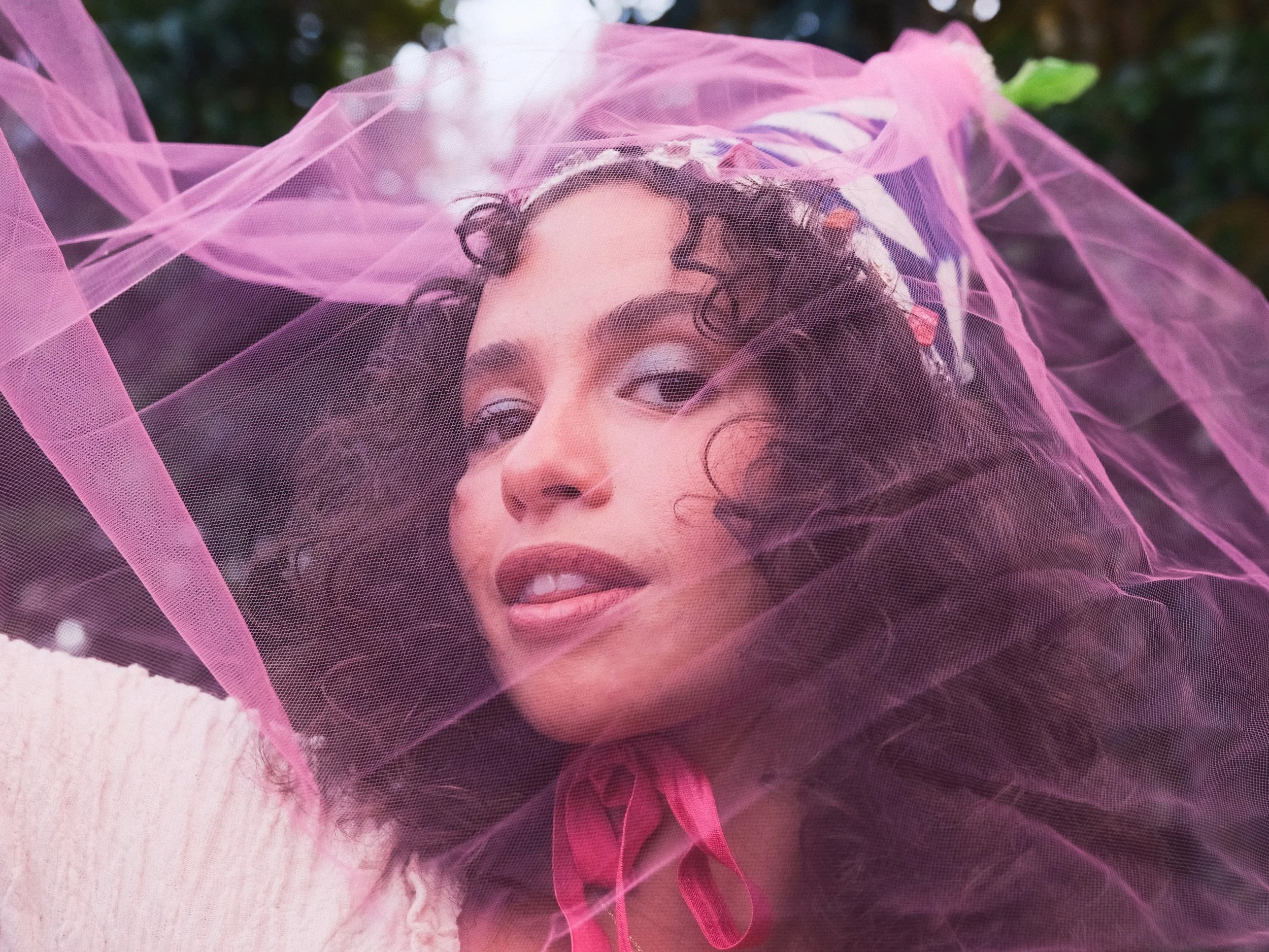 Close-up of a woman with curly hair wearing glasses and a pink veil, lying on a bed with a pink blanket, with an outdoor background.