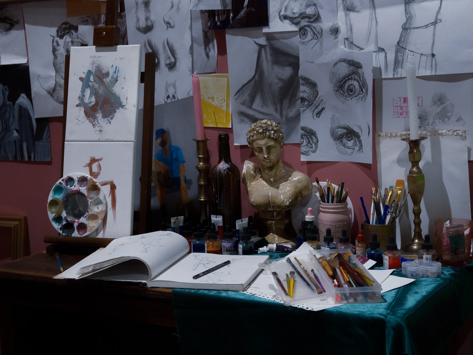 An artist's workspace with sketches of faces and eyes on the wall, an easel with an abstract painting, a palette, paint bottles, brushes, and drawing tools on the table, and a bust sculpture of a young man in the center.