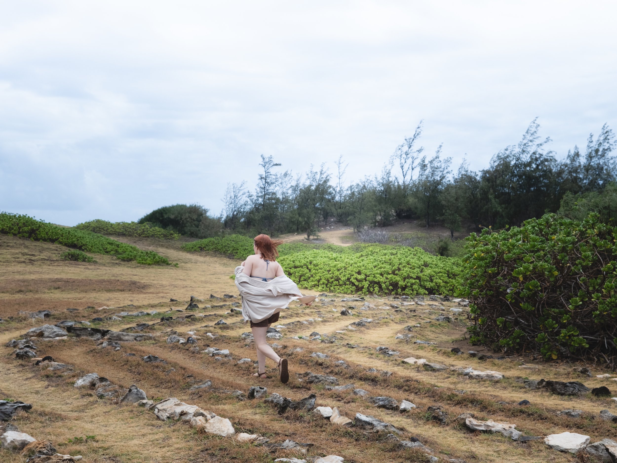 A woman with red hair sitting on a grassy field surrounded by rocks, with a backdrop of trees and a cloudy sky.