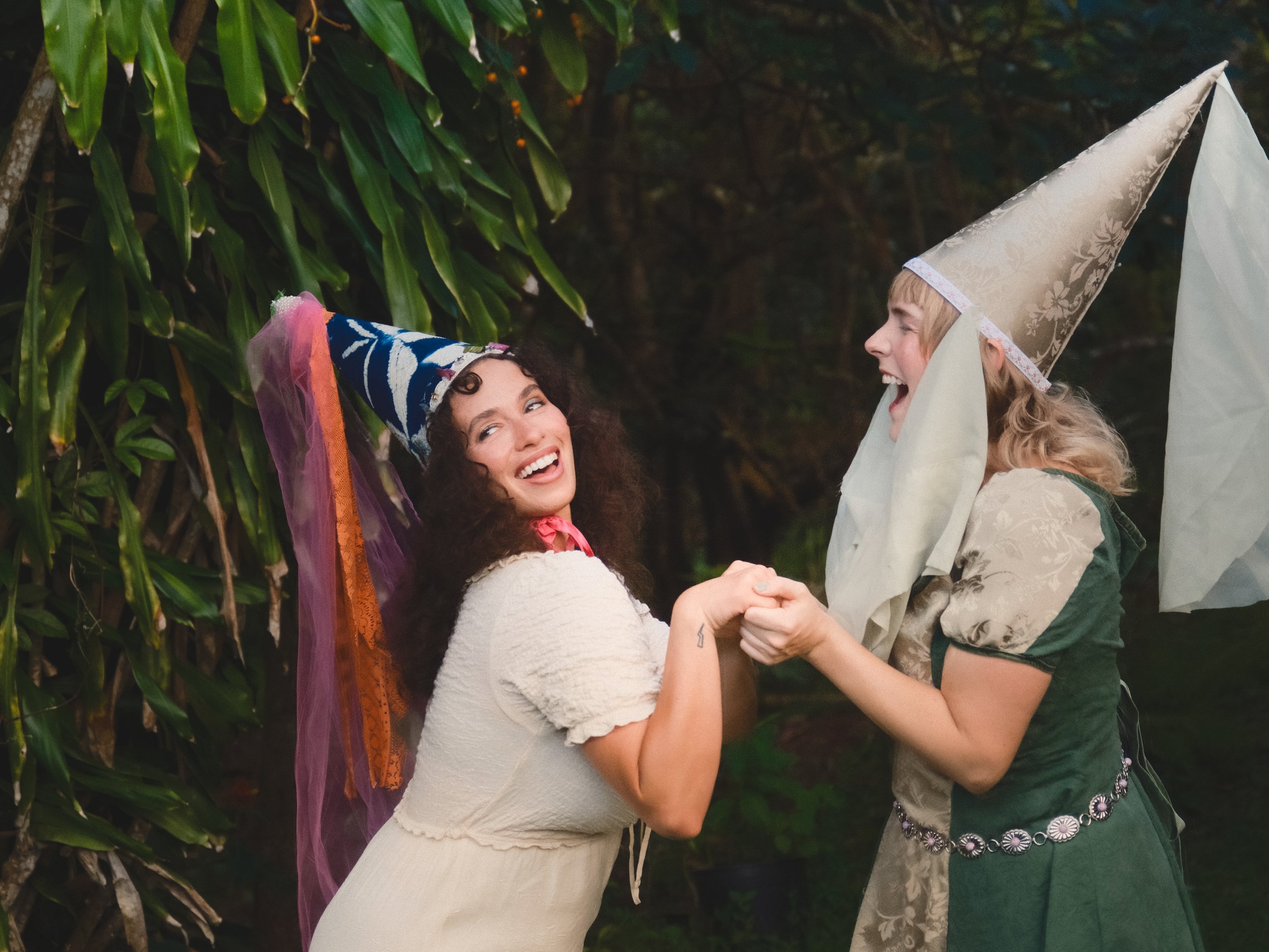Two women wearing pointed hats, smiling and holding hands outdoors near green leafy plants.