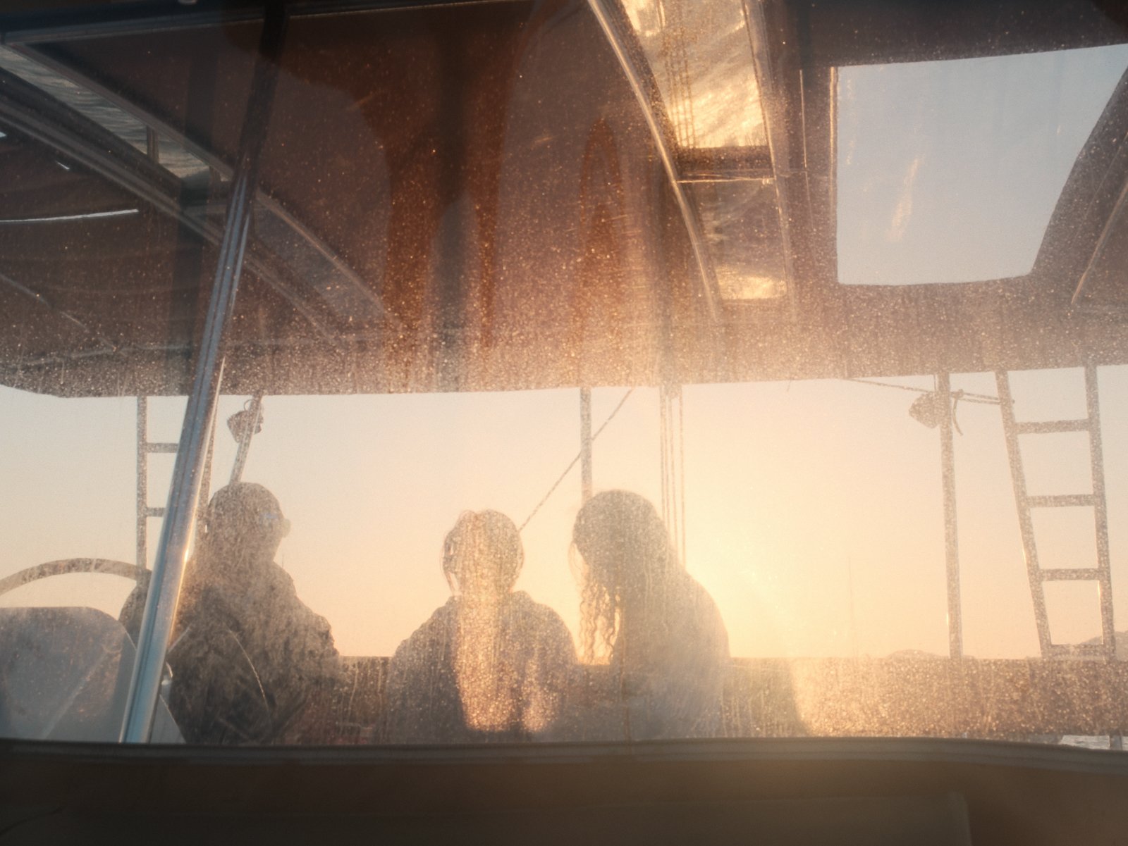 Three people sitting on a boat during sunset, viewed through a dusty window with ladder and metal framework visible.