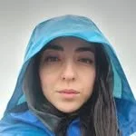 Close-up of a young woman wearing a blue waterproof hooded jacket, with dark hair and a neutral expression.