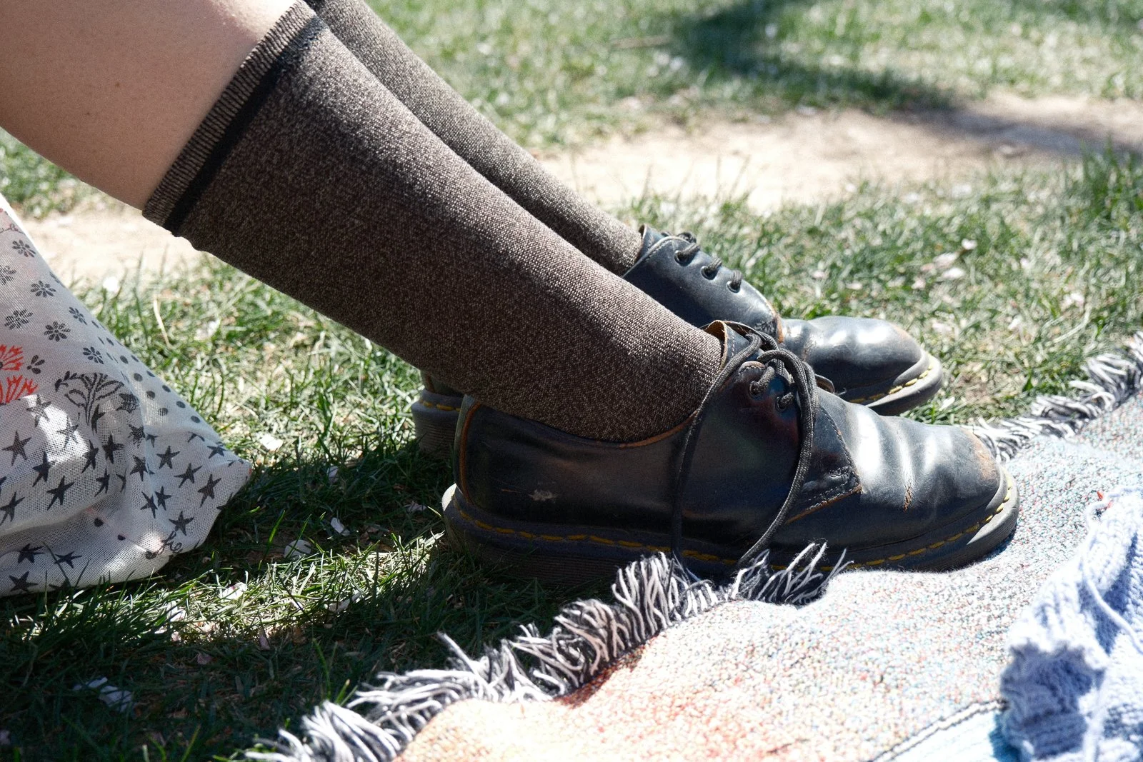 Close-up of black dress shoes and brown wool socks on a person's feet, outdoors on grass with a blanket nearby.