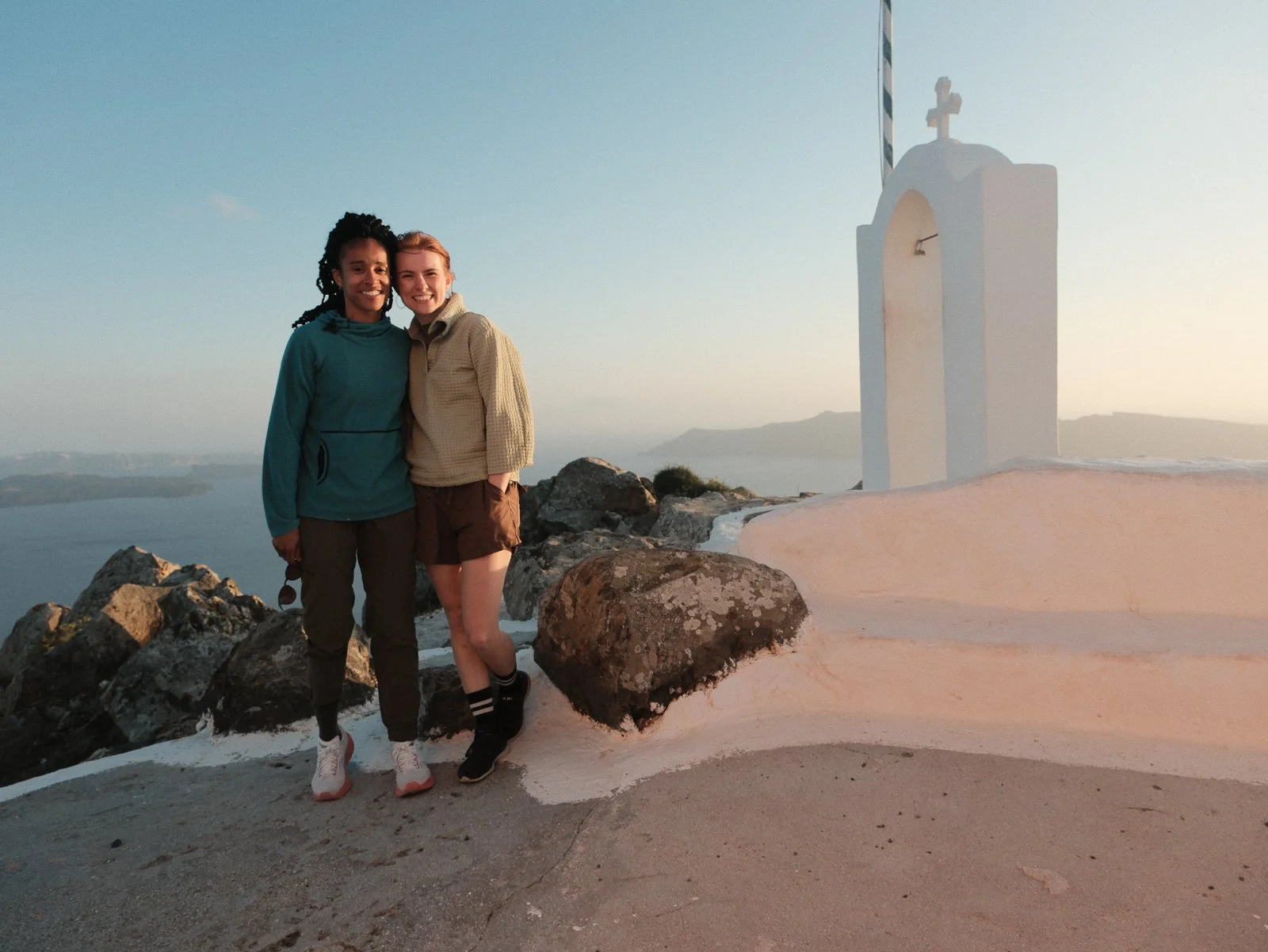 Two women standing on a rocky landscape at sunset, with a small white chapel and a cross in the background, overlooking the water and distant islands.
