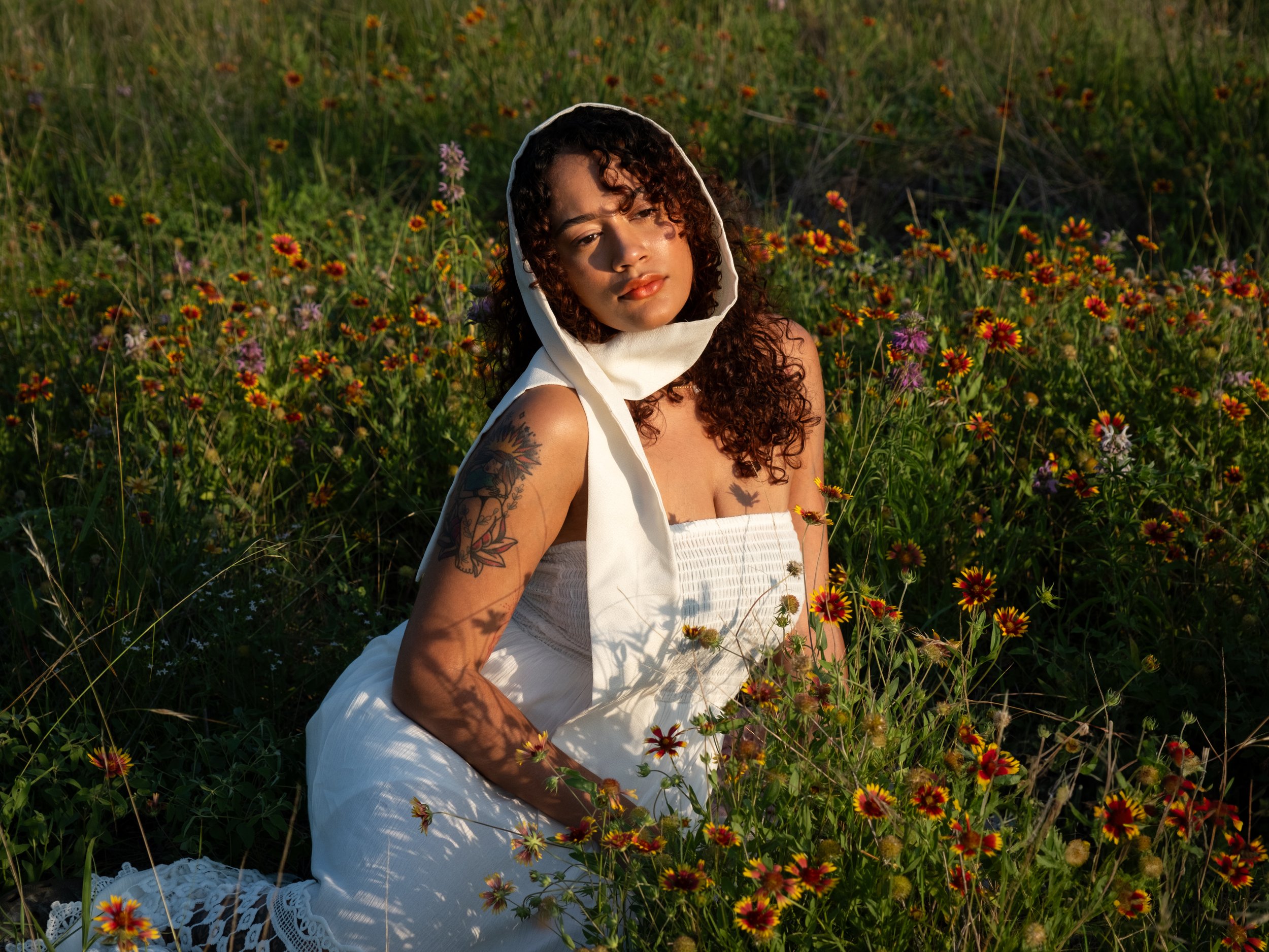 A woman with curly hair and tattoos wearing a white dress with a hood, sitting in a field of colorful wildflowers, looking peacefully at the camera.