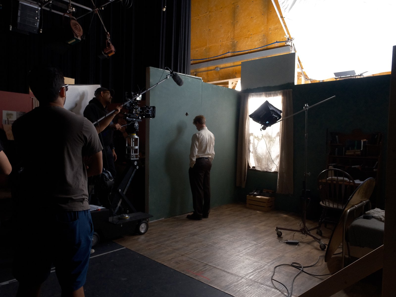 Behind the scenes of a film or video shoot, crew members adjusting equipment, a man standing in front of a green screen, and filming lights in a room with curtains and furniture.