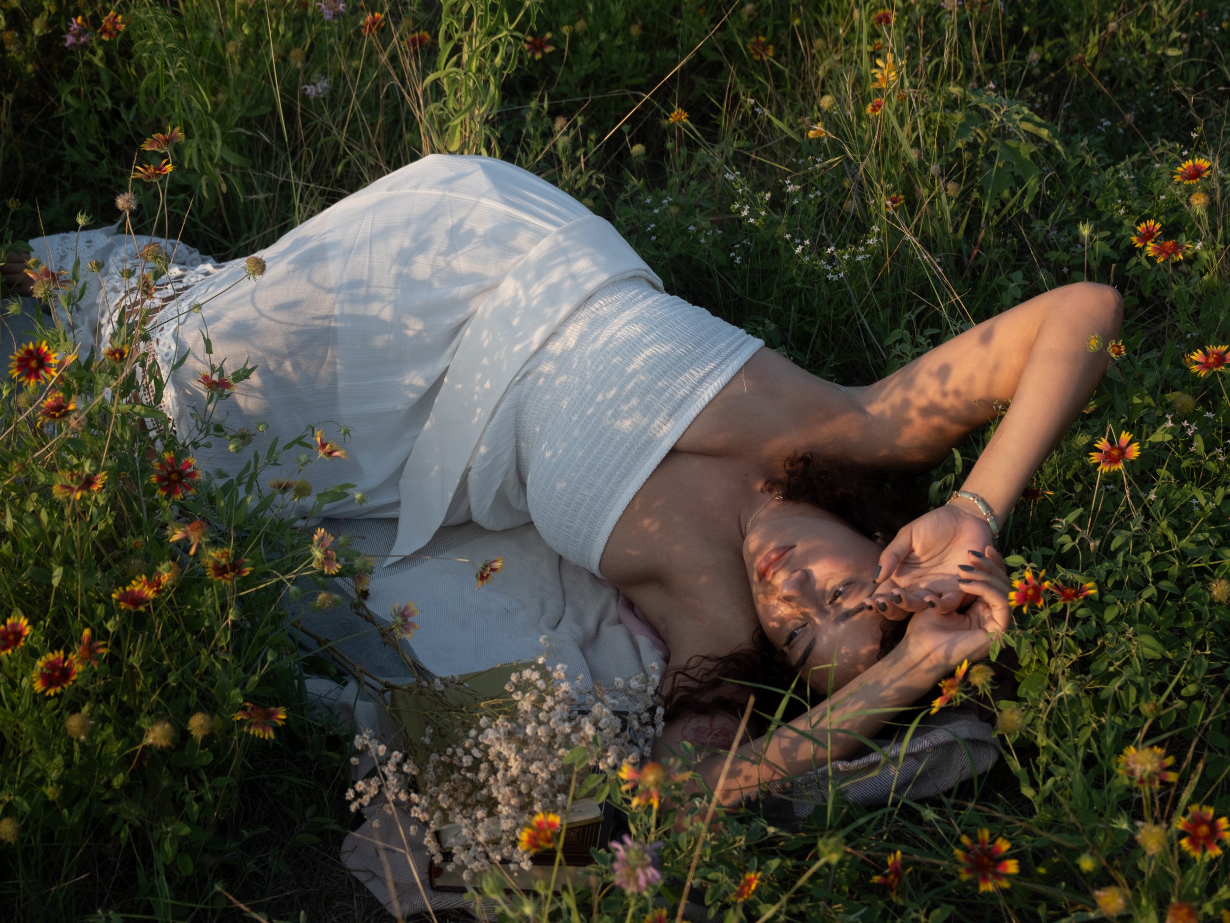 Person lying down on the grass among wildflowers, wearing a white dress and a bracelet
