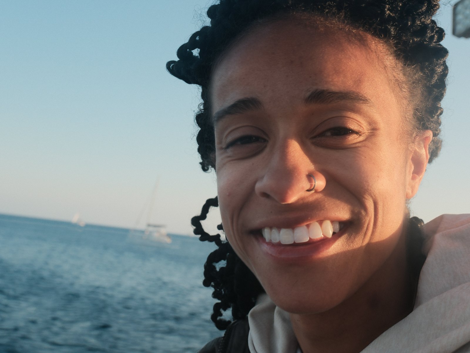 Close-up portrait of a young woman with a big smile, short curly hair, and a nose ring, enjoying a seaside view with sailboats in the background.