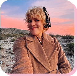 A woman with blonde curly hair wearing headphones and a brown coat, smiling outdoors during sunset with a pink and blue sky and a desert landscape in the background.