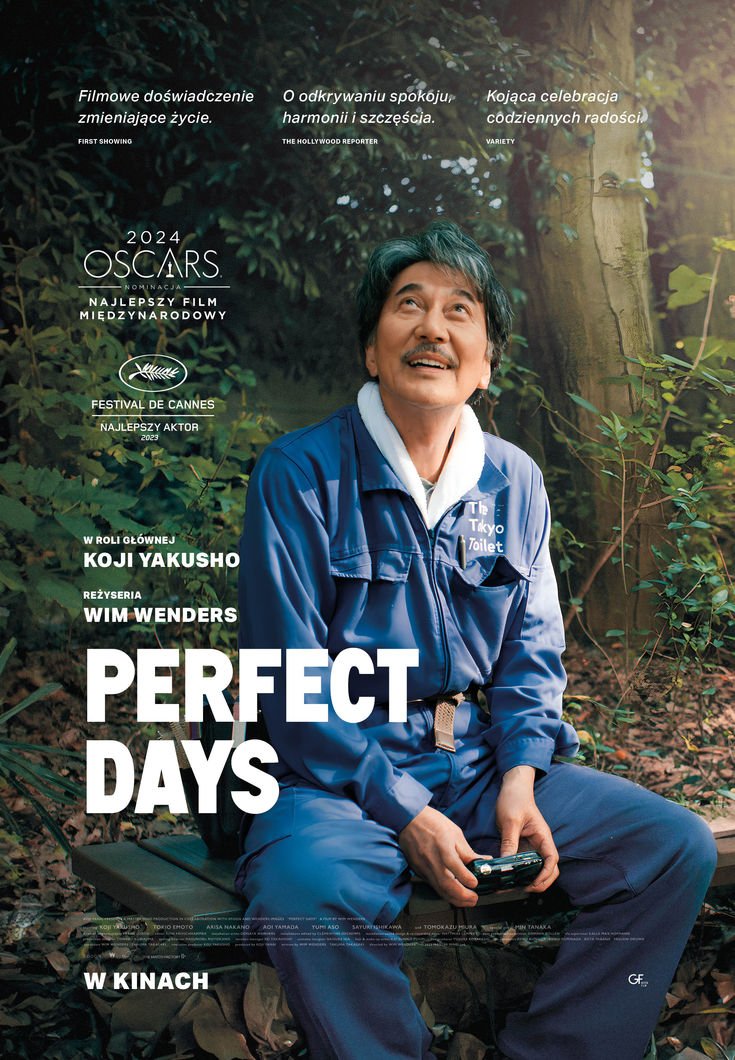 A man dressed in a blue jacket and pants sitting on a bench in a wooded area, holding a remote control device, smiling while looking upwards.