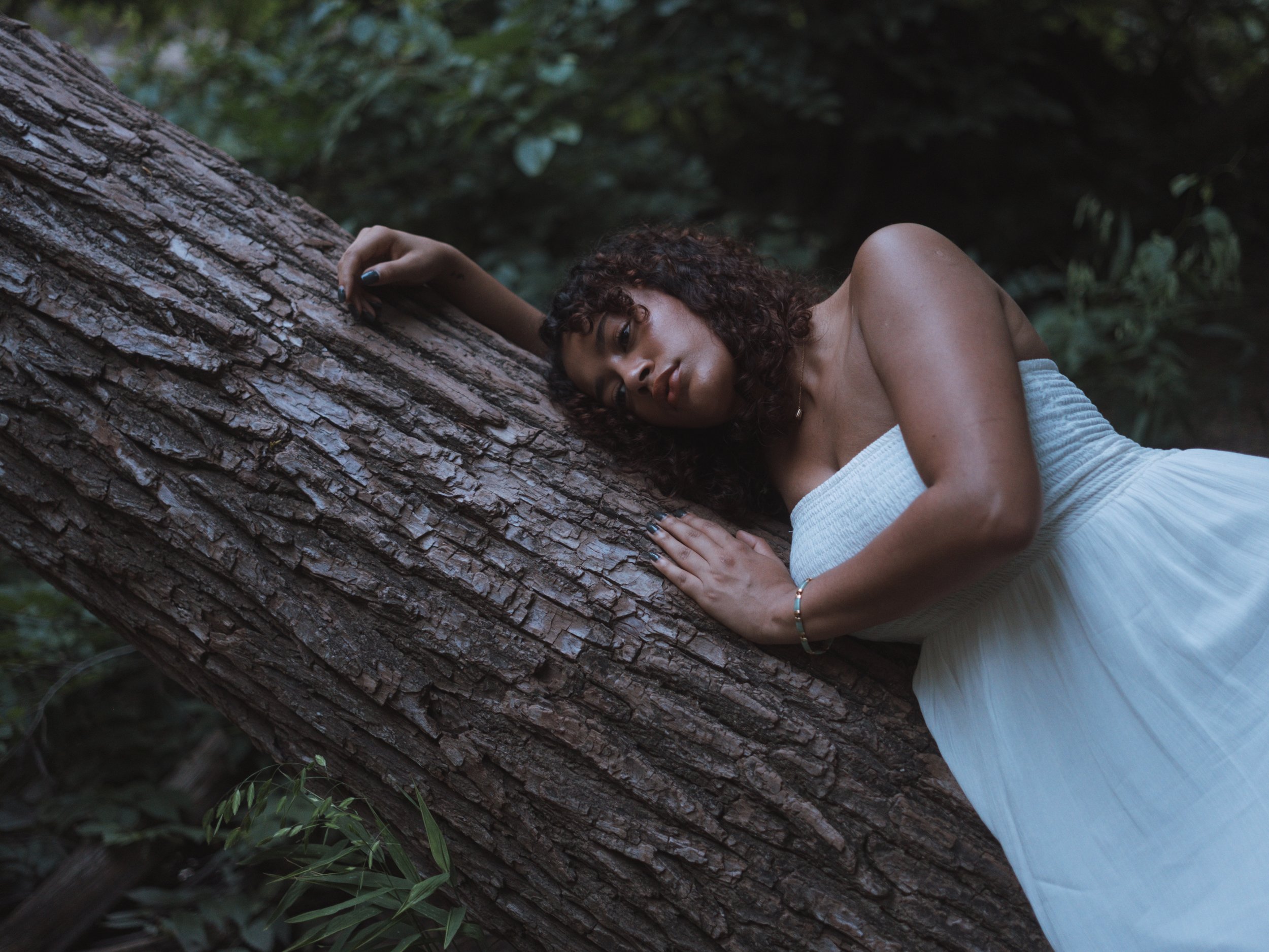 A woman with curly hair lying on a fallen tree trunk in a forest, wearing a white strapless dress, with her head resting on the tree and looking relaxed.