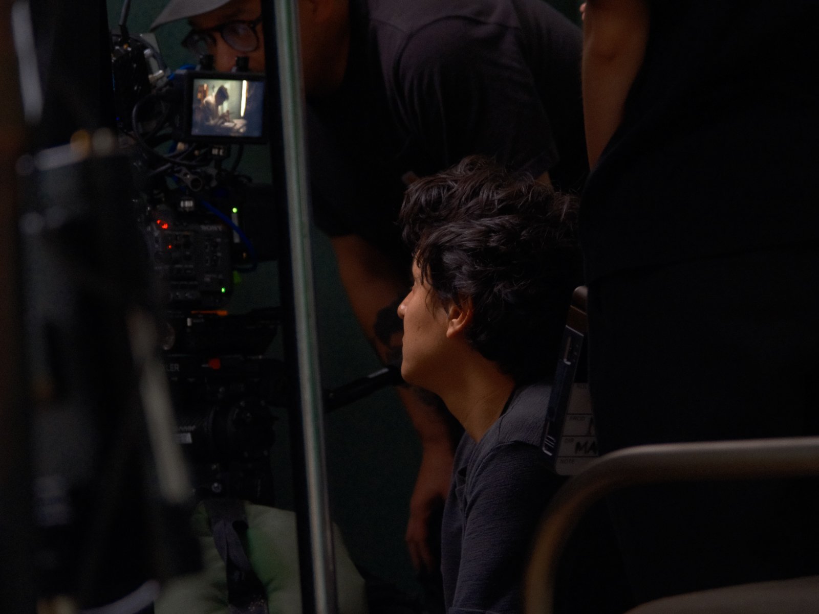 A man with curly hair is sitting in front of a video camera setup, with two crew members standing around him in a dimly lit setting, possibly on a film or TV set.