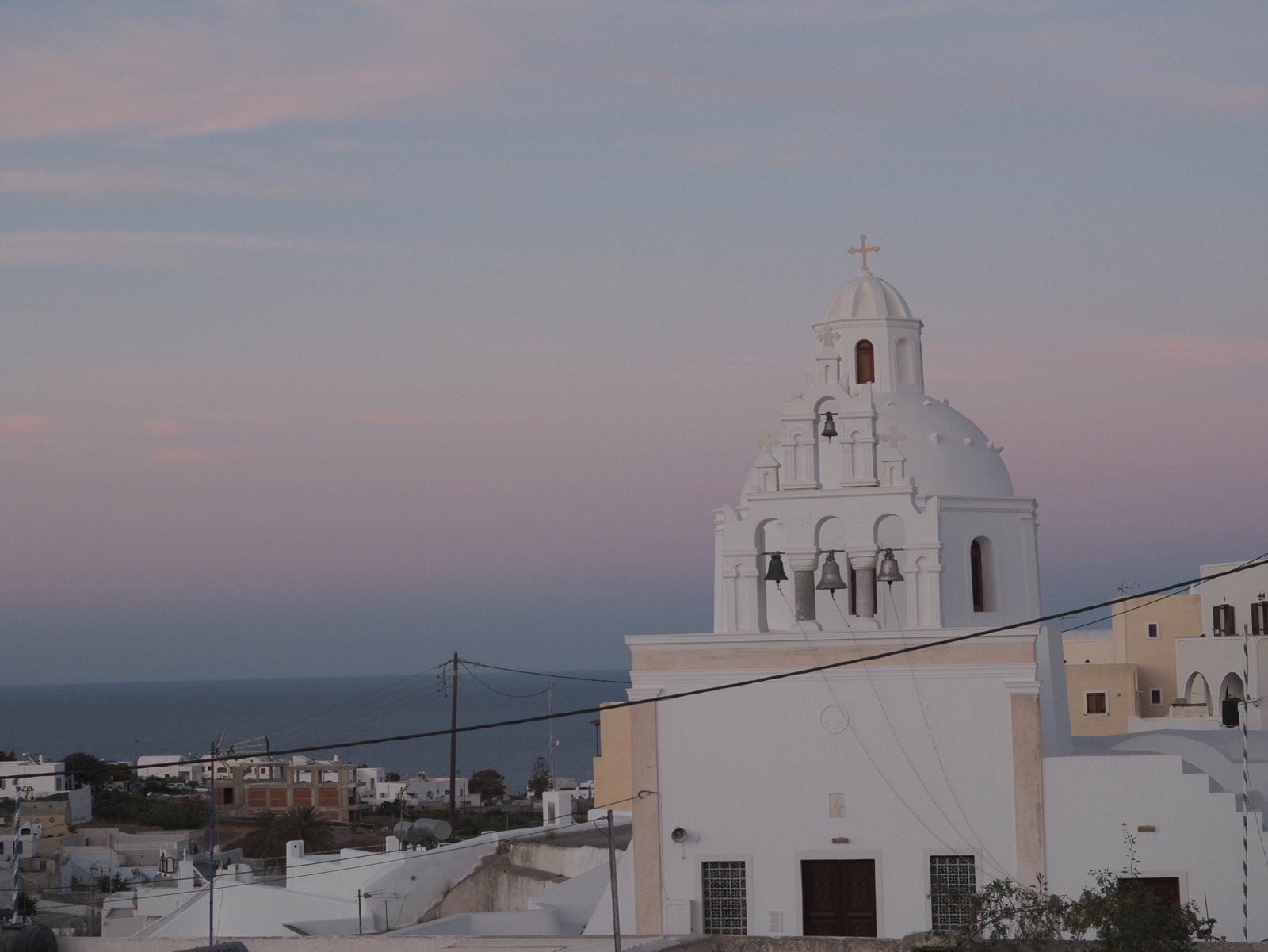 A white church with a bell tower topped with a cross, set against a dusky sky, with surrounding buildings and power lines in the foreground.