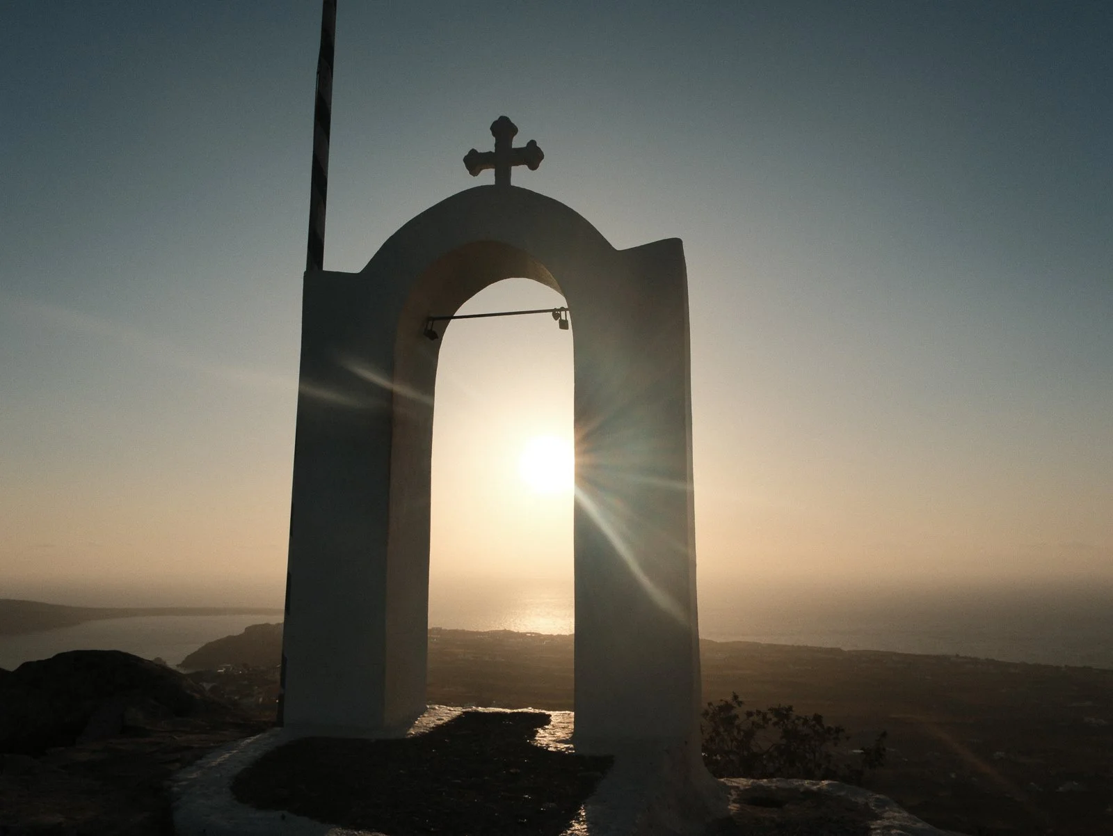 A white bell tower with a cross on top, silhouetted against a sunset sky with visible sun rays, overlooking an ocean and coastline.