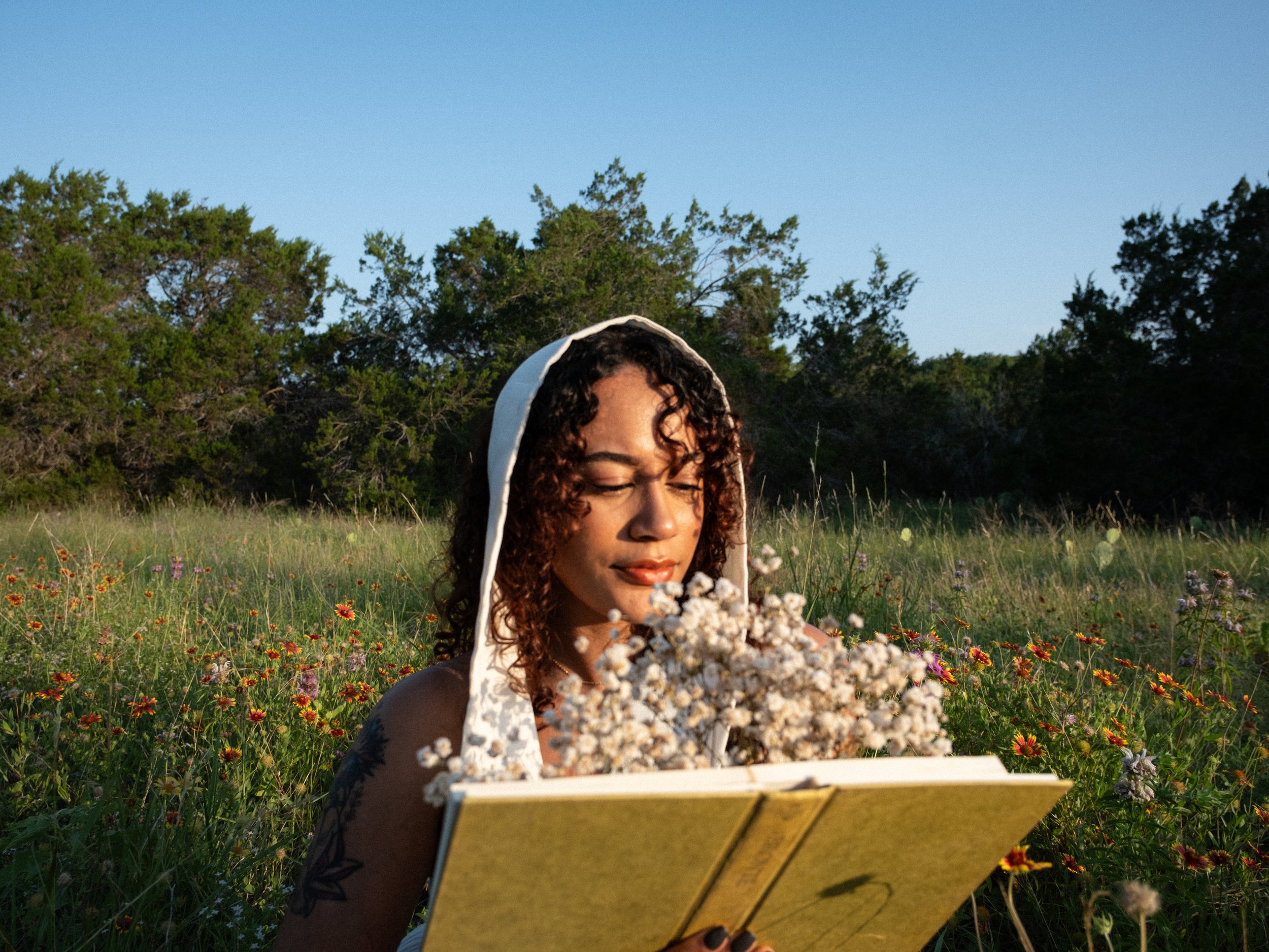 A woman with curly hair and a white hood on her head is sitting in a field of flowers and reading a yellow book.
