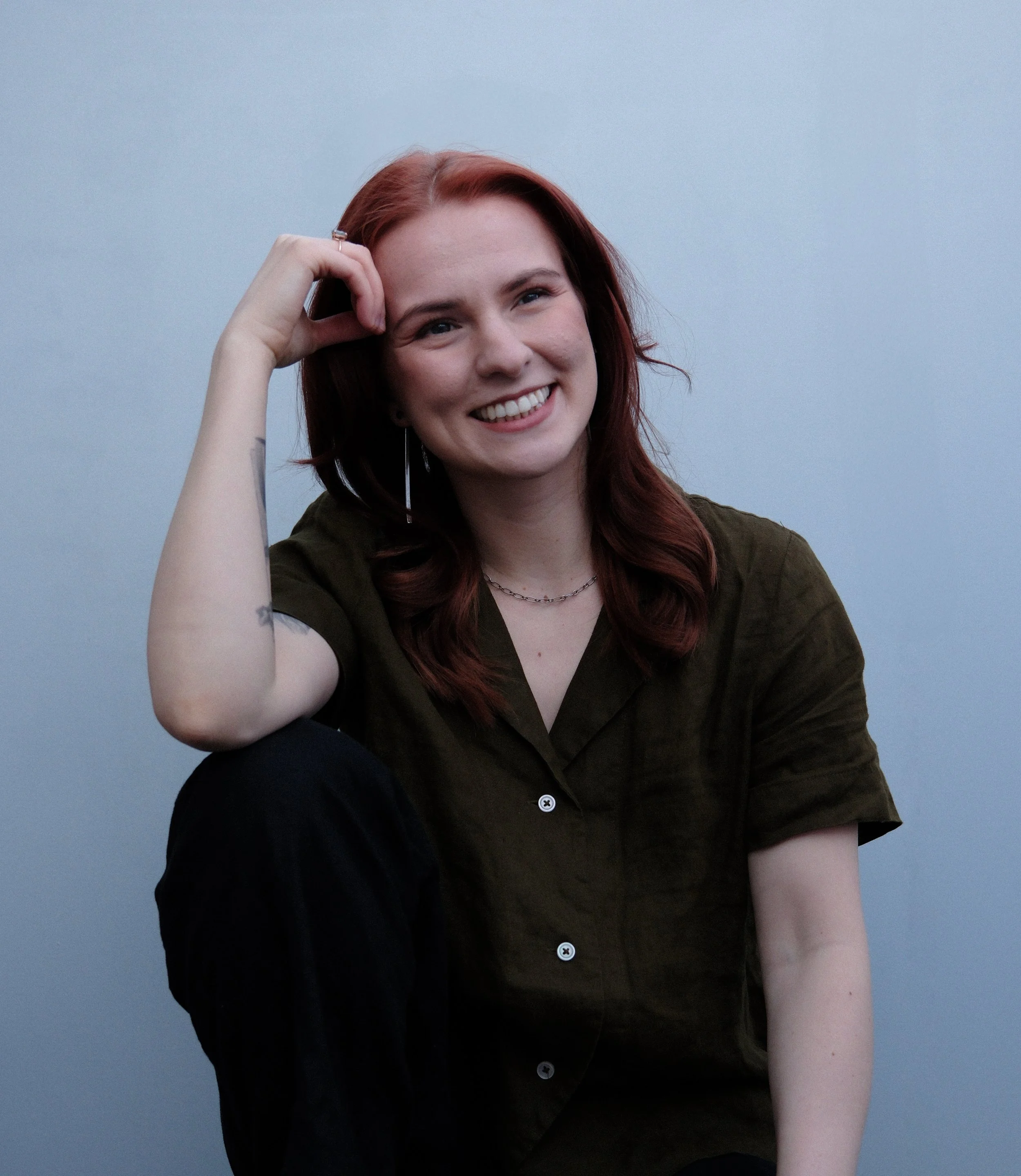 A smiling woman with reddish-brown hair, earrings, and a necklace, sitting with her arm resting on her knee against a plain blue background.
