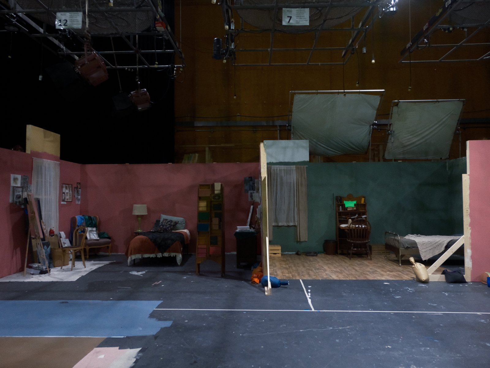 Stage set with furniture and props for a play, including chairs, a bed, a lamp, and a bookshelf, with painted walls and overhead stage lighting.