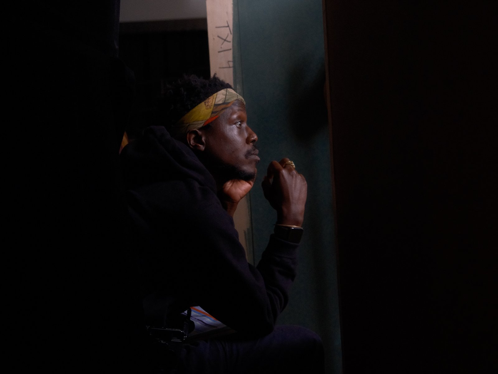 A man with dark skin, wearing a patterned headwrap, sitting in a dimly lit room, resting his chin on his hand and looking intently to the side.