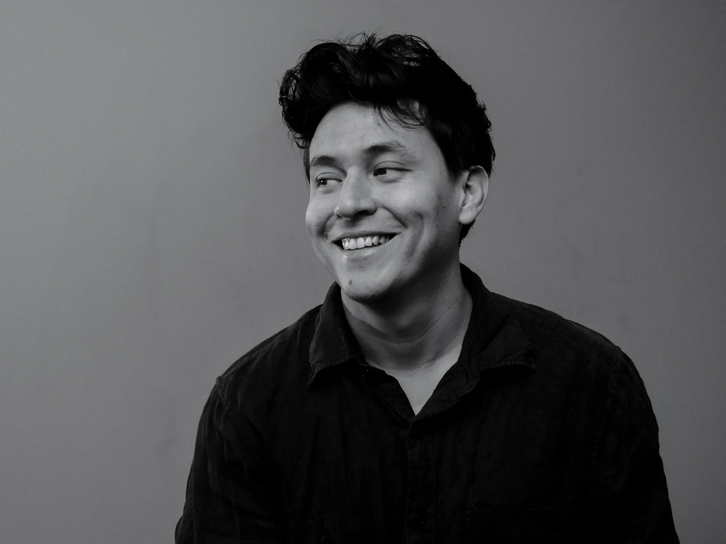 A smiling young man with dark, messy hair wearing a dark shirt, looking to the side against a plain background.