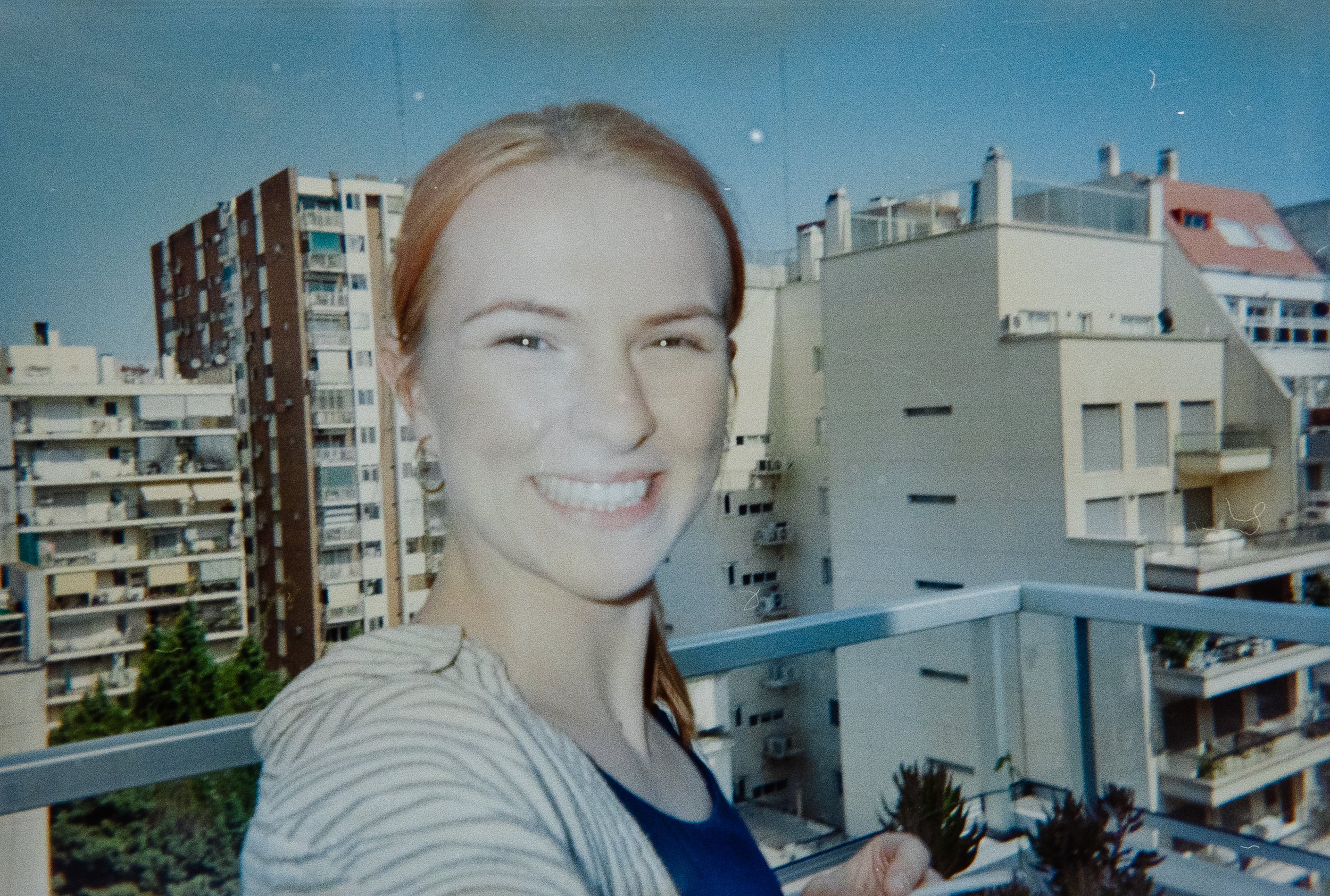 A smiling woman with reddish hair takes a selfie on a balcony with tall apartment buildings in the background under a clear blue sky.
