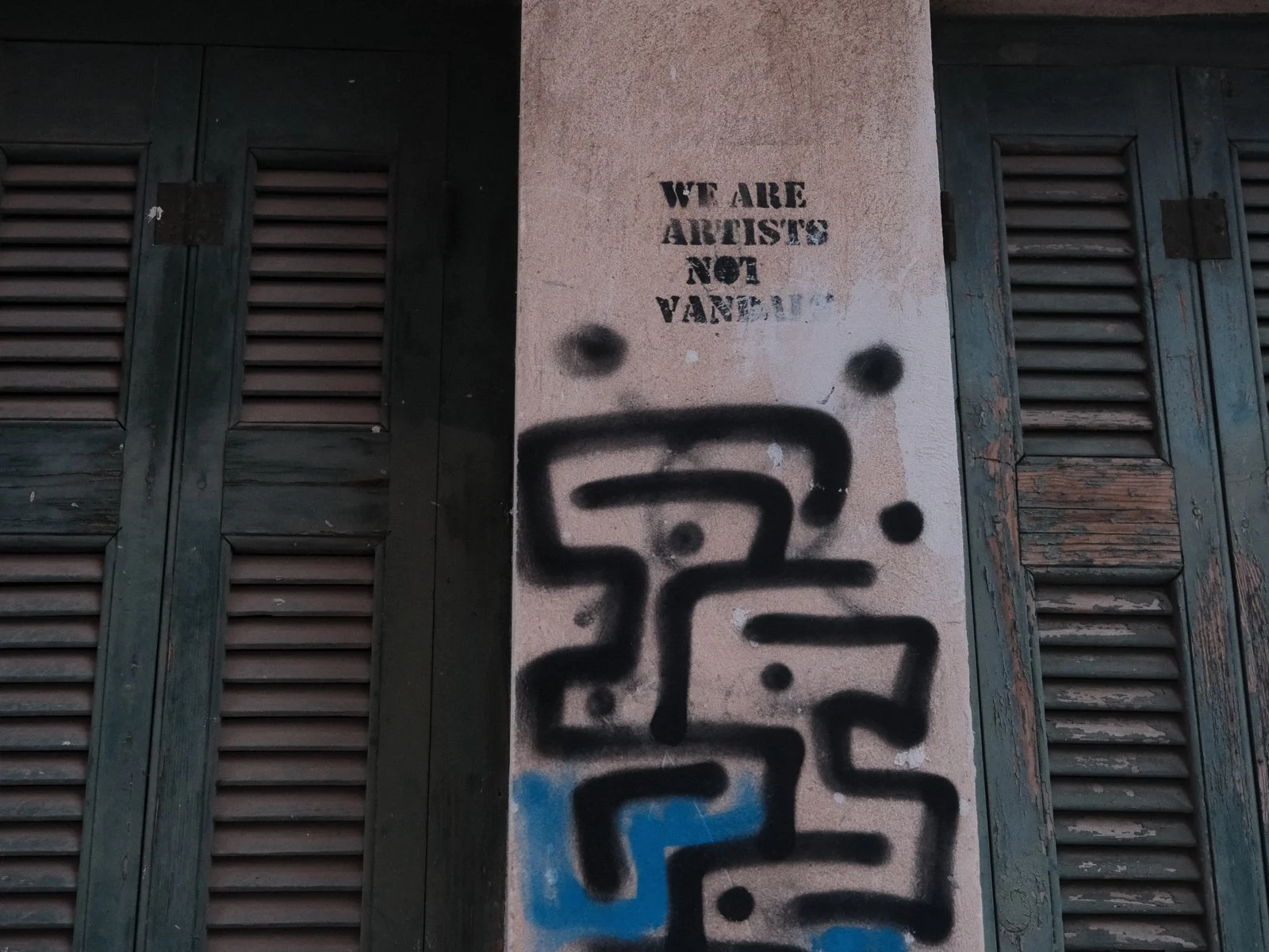 A wall with a stencil graffiti message reading 'We are artists not vandals' and black spray-painted graffiti that appears to spell 'peace' on a pinkish-beige surface near window shutters.
