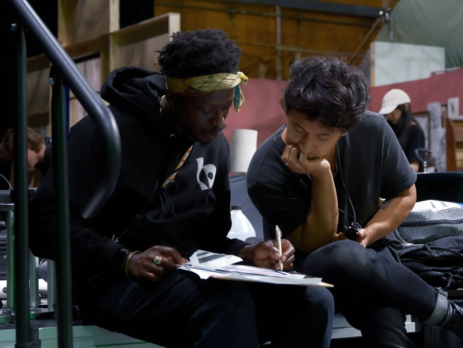 Two women sit closely together, engaged in conversation and working on a document or notebook. One woman wears a yellow headband and a black hoodie, while the other has short curly hair and wears a dark T-shirt. The setting appears to be an indoor sp