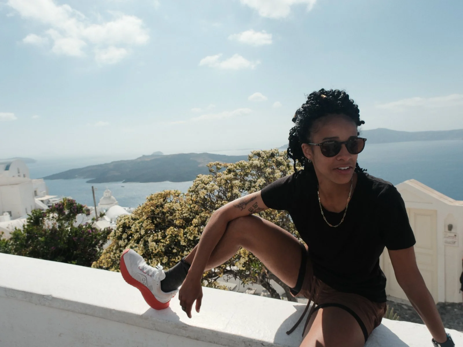 A person with curly hair and sunglasses sitting on a white wall in a scenic coastal area with white buildings, flowers, and a view of the ocean and distant islands.