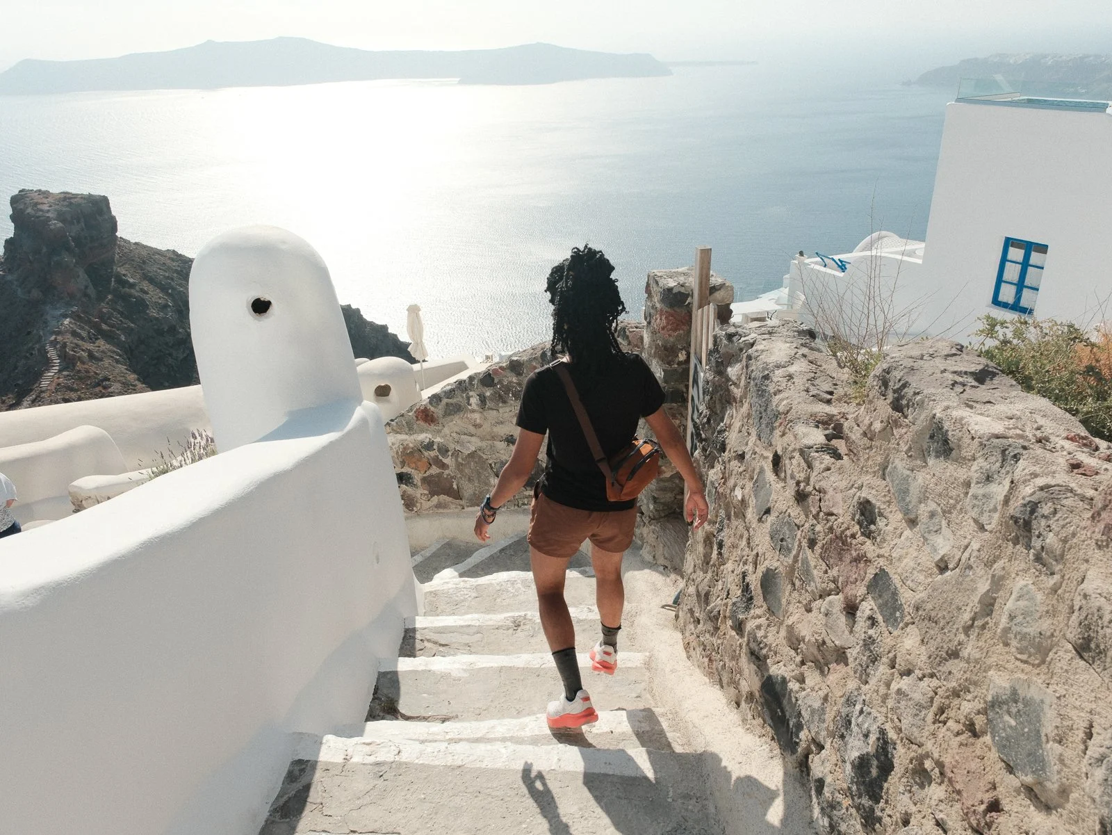 A person with black curly hair, wearing a black shirt, brown shorts, and white sneakers with orange soles, walking down white stairs toward the sea with a rocky wall on the right and white architecture on the left. The background features a calm body