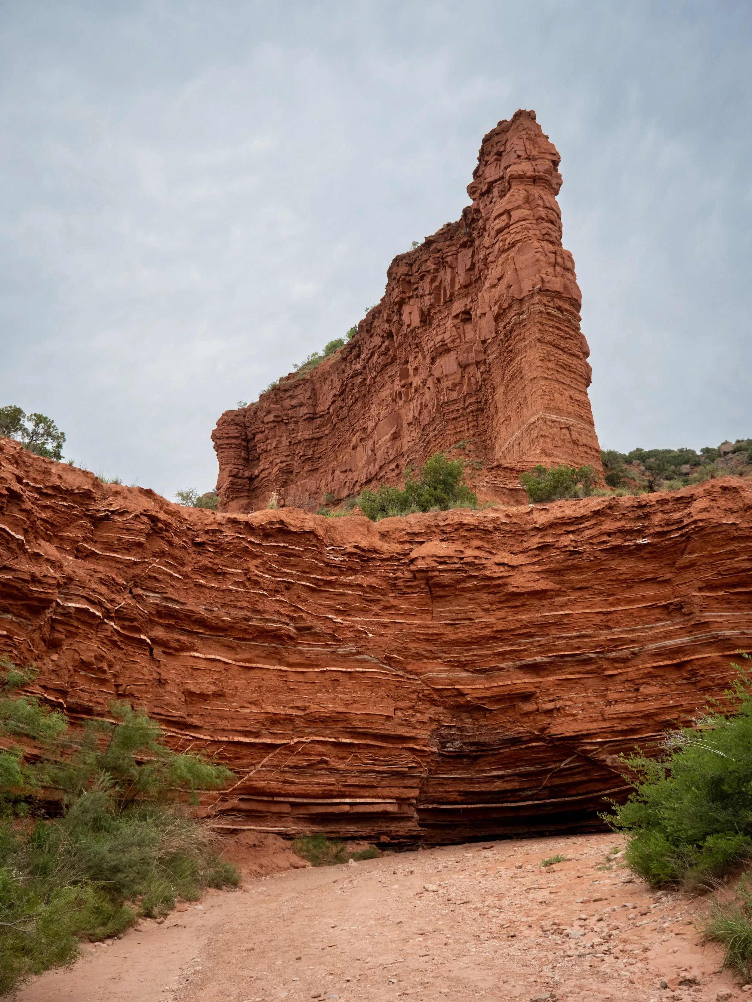 Red rock formations with layered cliffs and a sandy dirt trail in a desert landscape, green bushes at the base, overcast sky overhead.