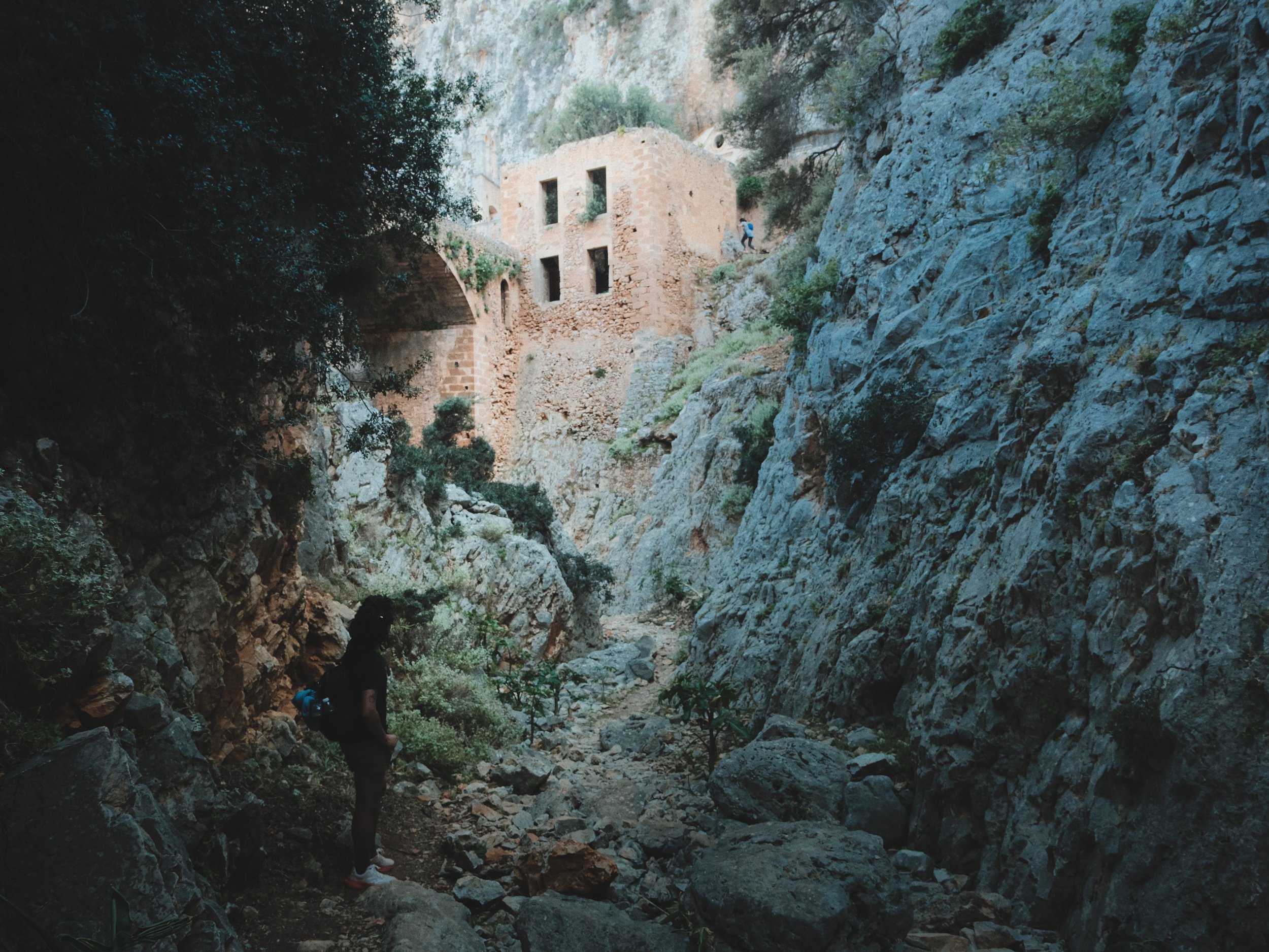 Hikers walking through a rocky canyon with an old stone building built into the cliffside and surrounded by trees.