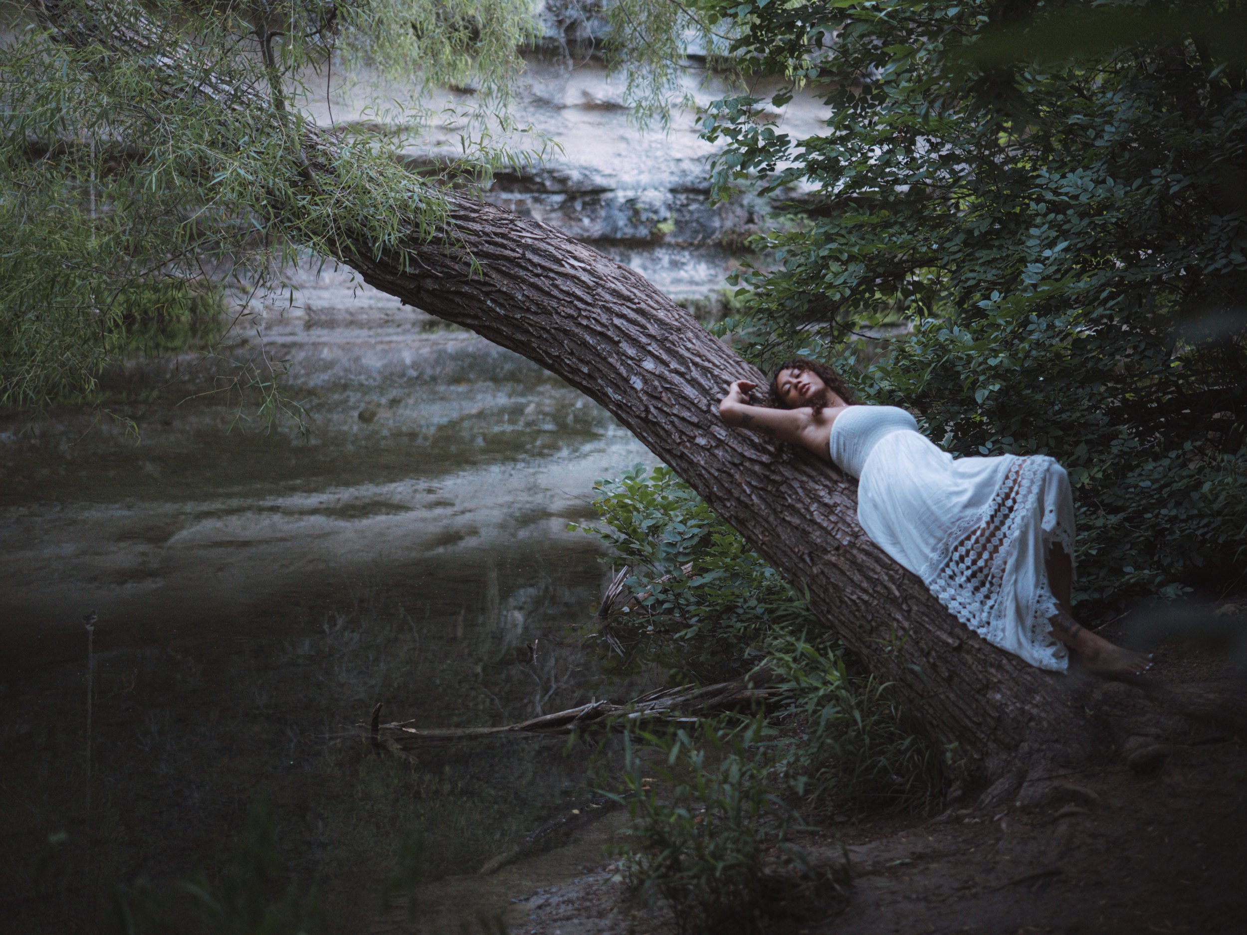 A woman in a white dress lying on a fallen tree trunk across a river, surrounded by lush green trees and bushes.