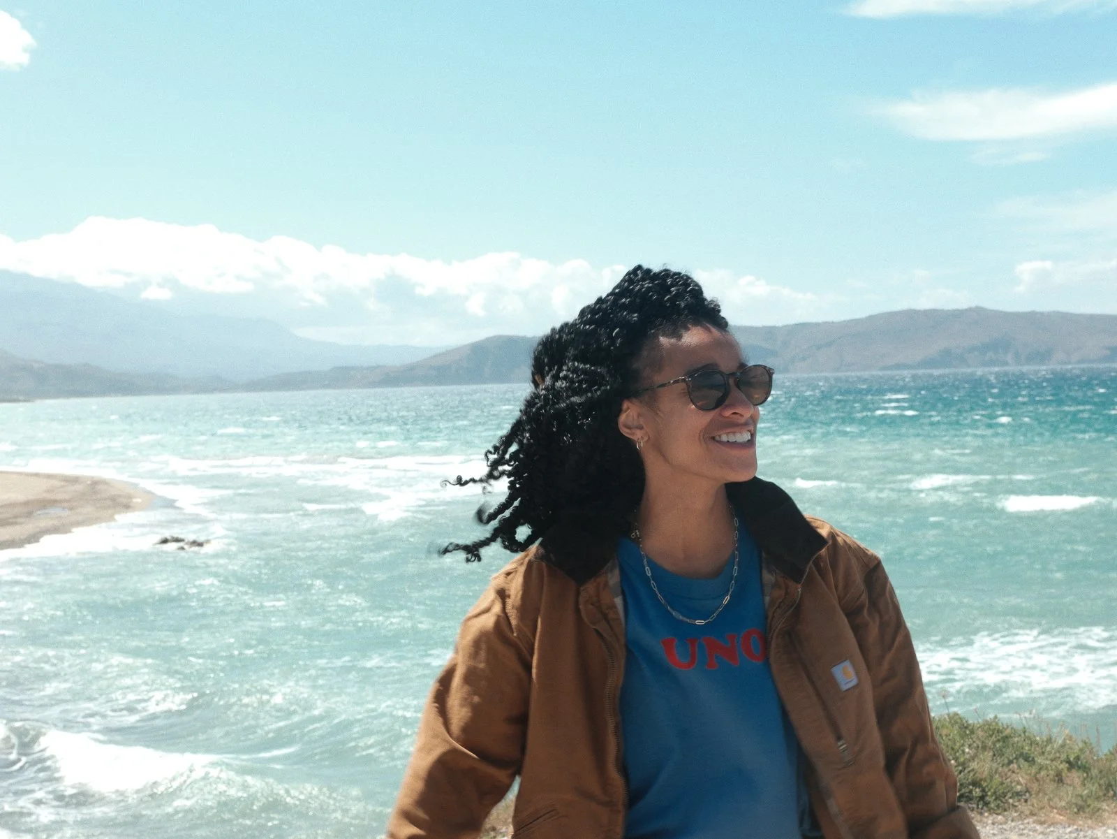 Smiling woman with curly hair wearing sunglasses, a brown jacket, and a blue T-shirt stands near a body of water with hills and clouds in the background.