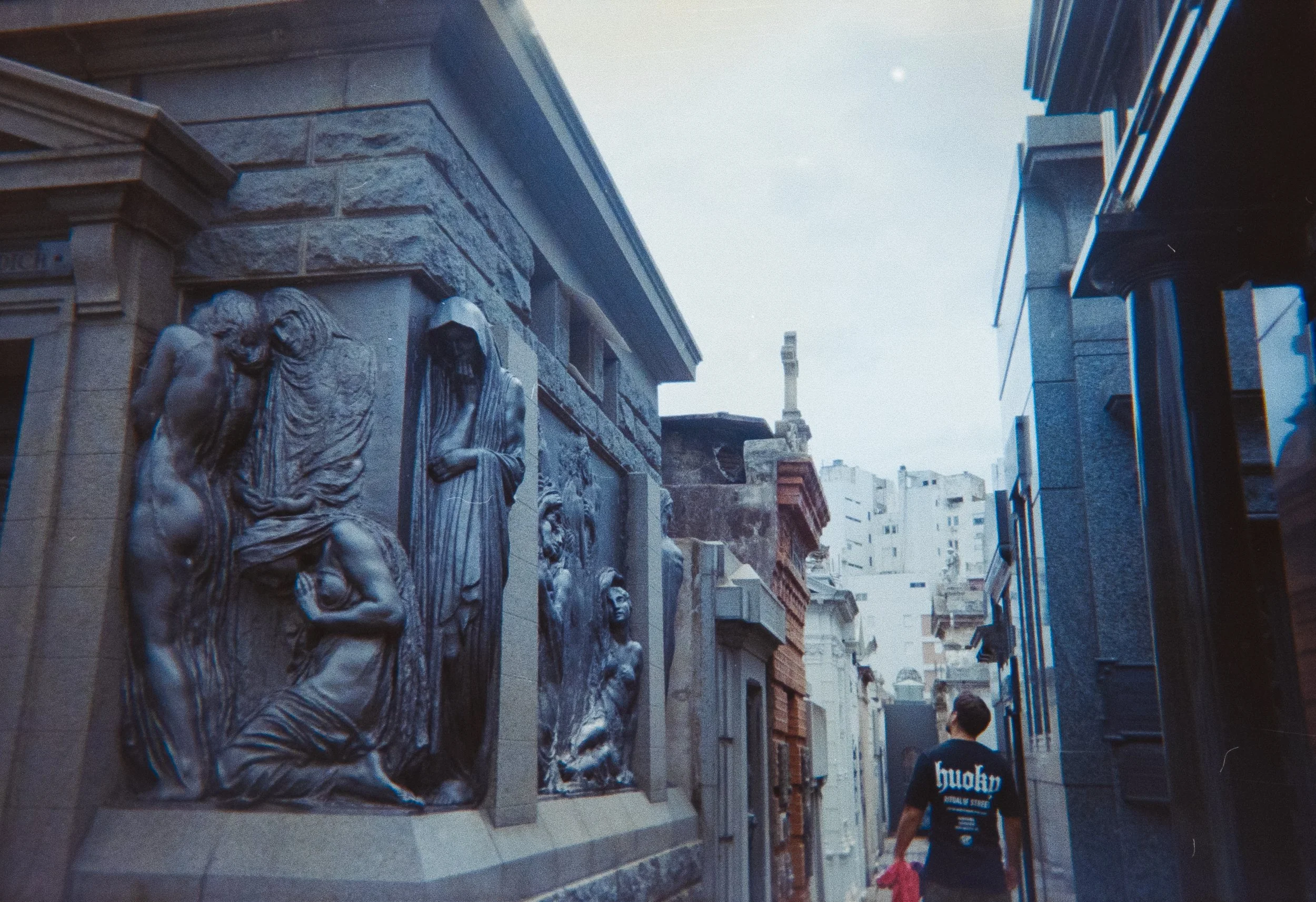 People walking past a row of religious statues and relief sculptures on a building's exterior wall in an urban area.