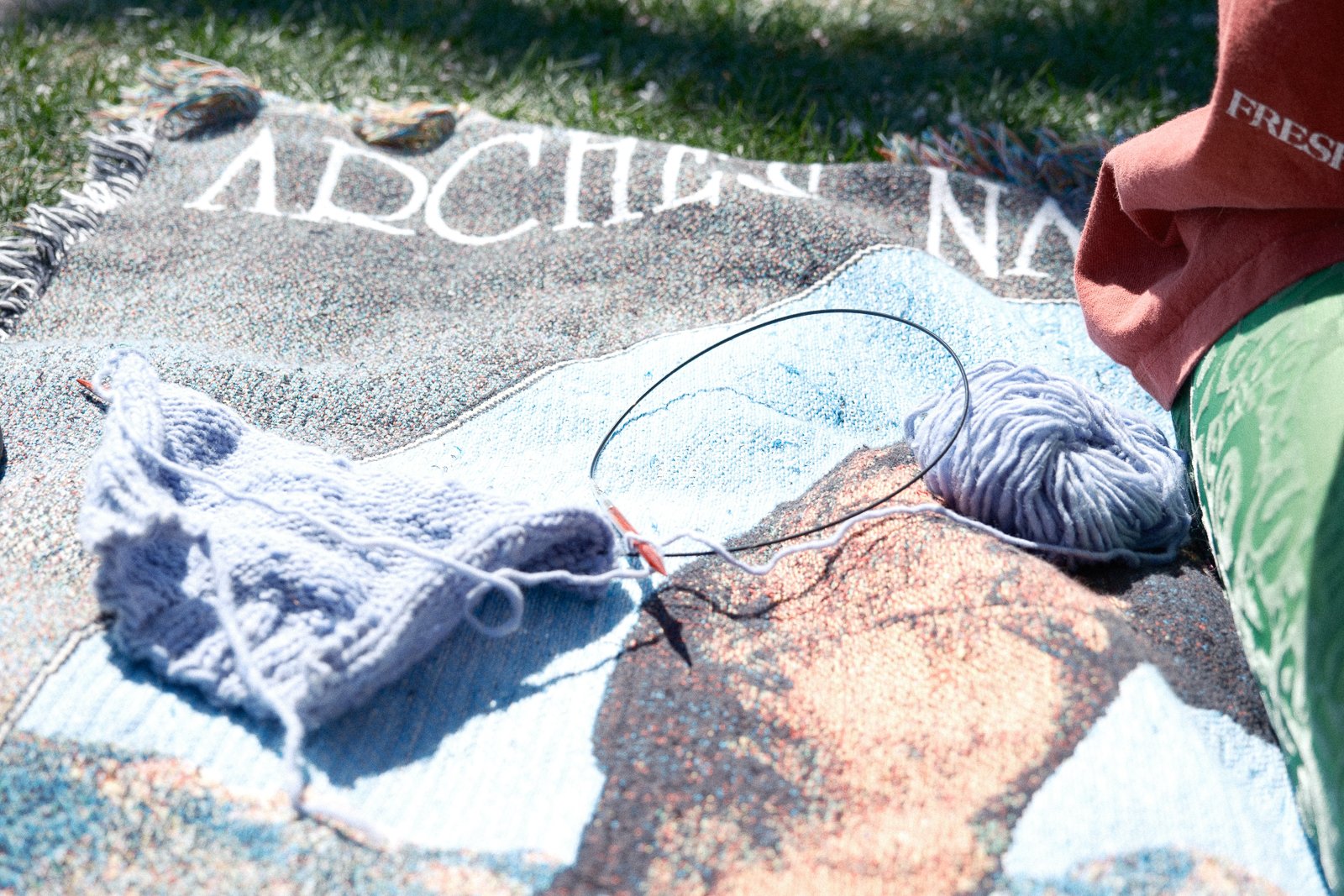 A close-up of a colorful beach towel on the sand with knitting supplies, including white yarn, knitting needles, and a ball of yarn, surrounded by grass and a person's arm and clothing.
